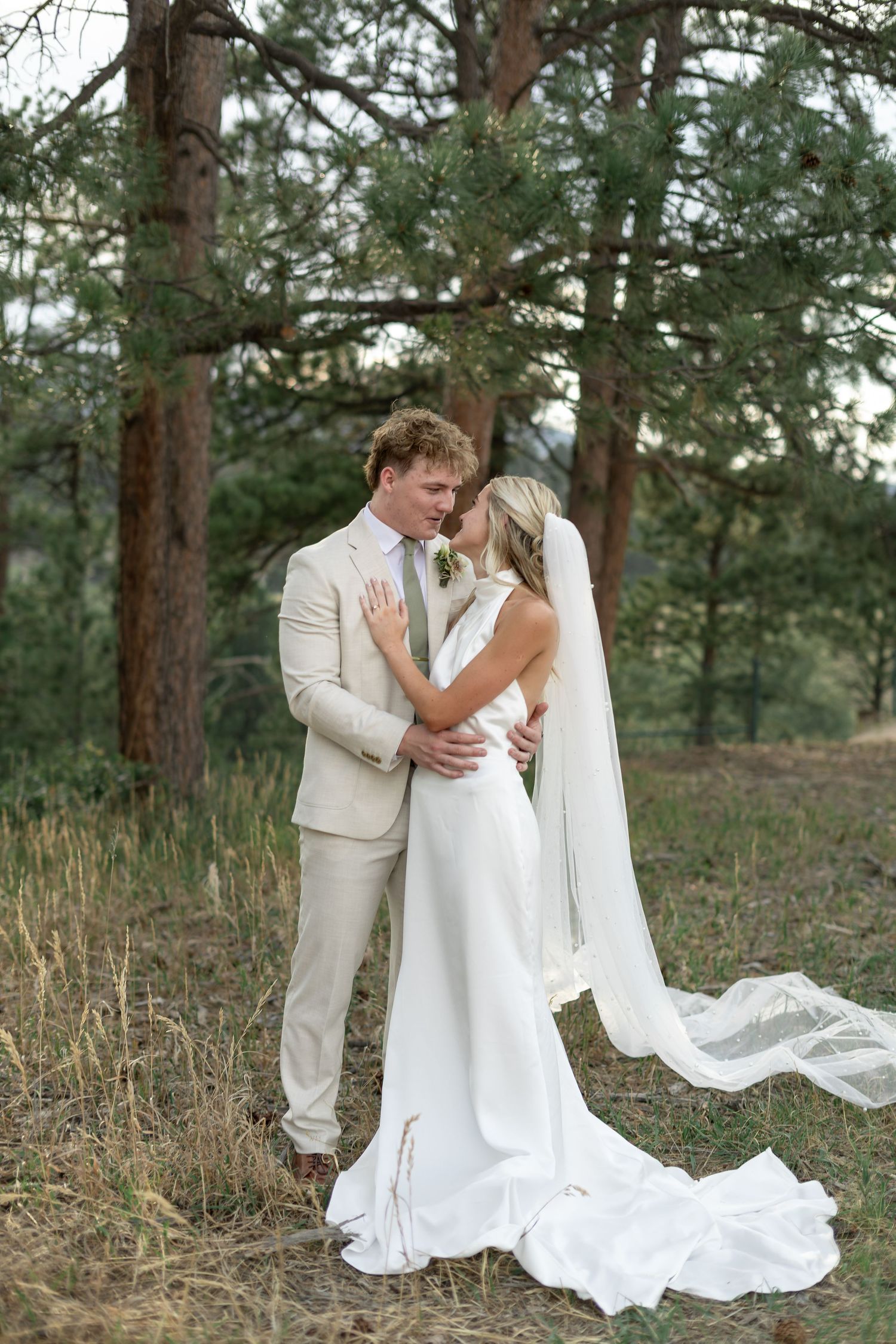 A bride and groom share a romantic moment in a forest, she in a flowing white gown with veil, he in a tan suit.