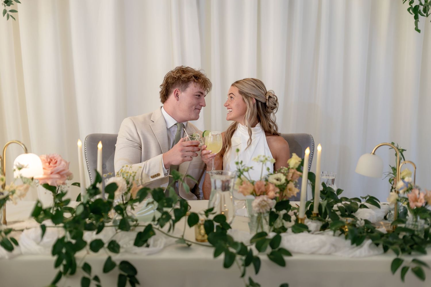 Bride and groom toasting with drinks at elegant floral wedding sweetheart table with candles and greenery.