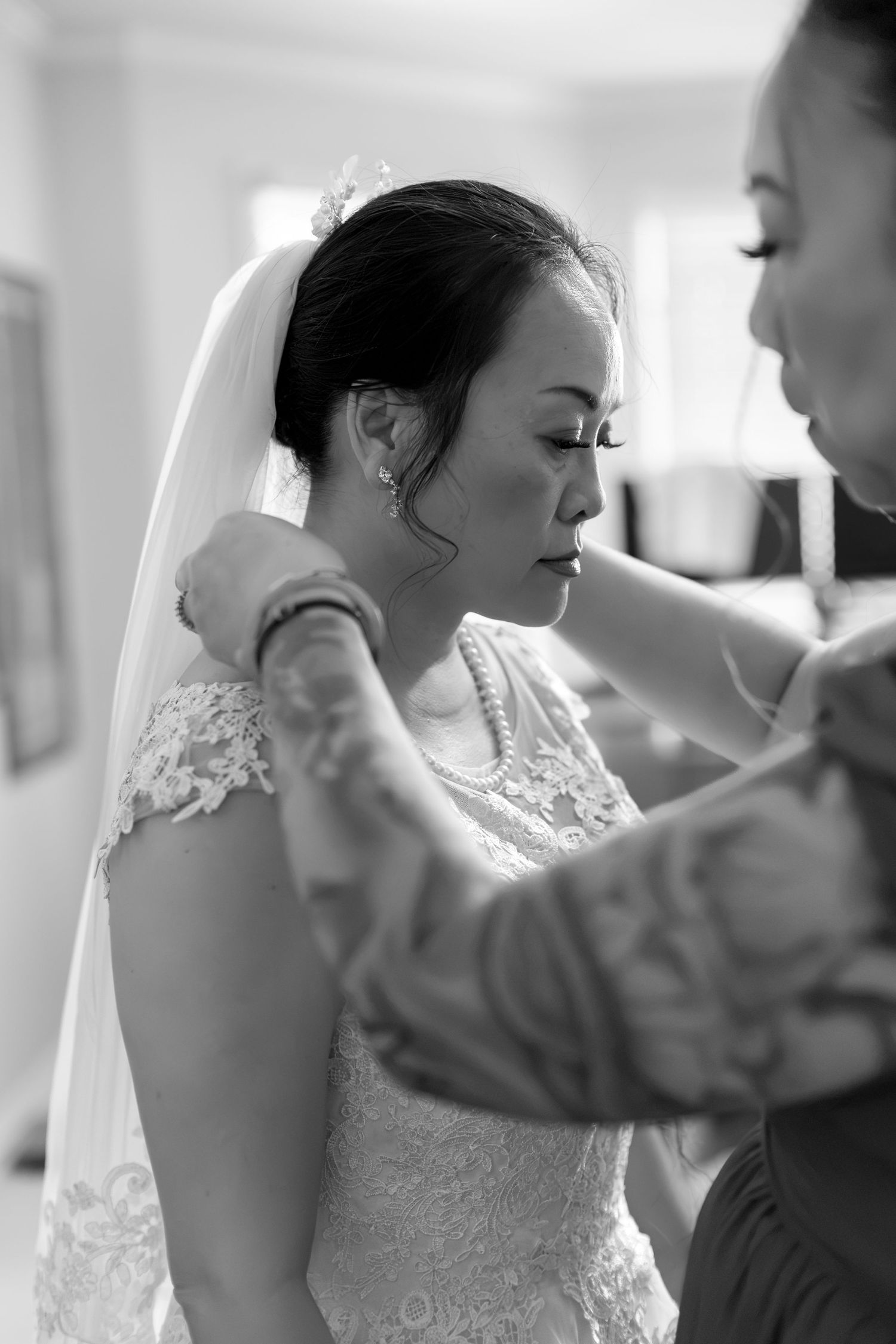Bride in lace wedding dress and veil being helped by bridesmaid in black and white photo.