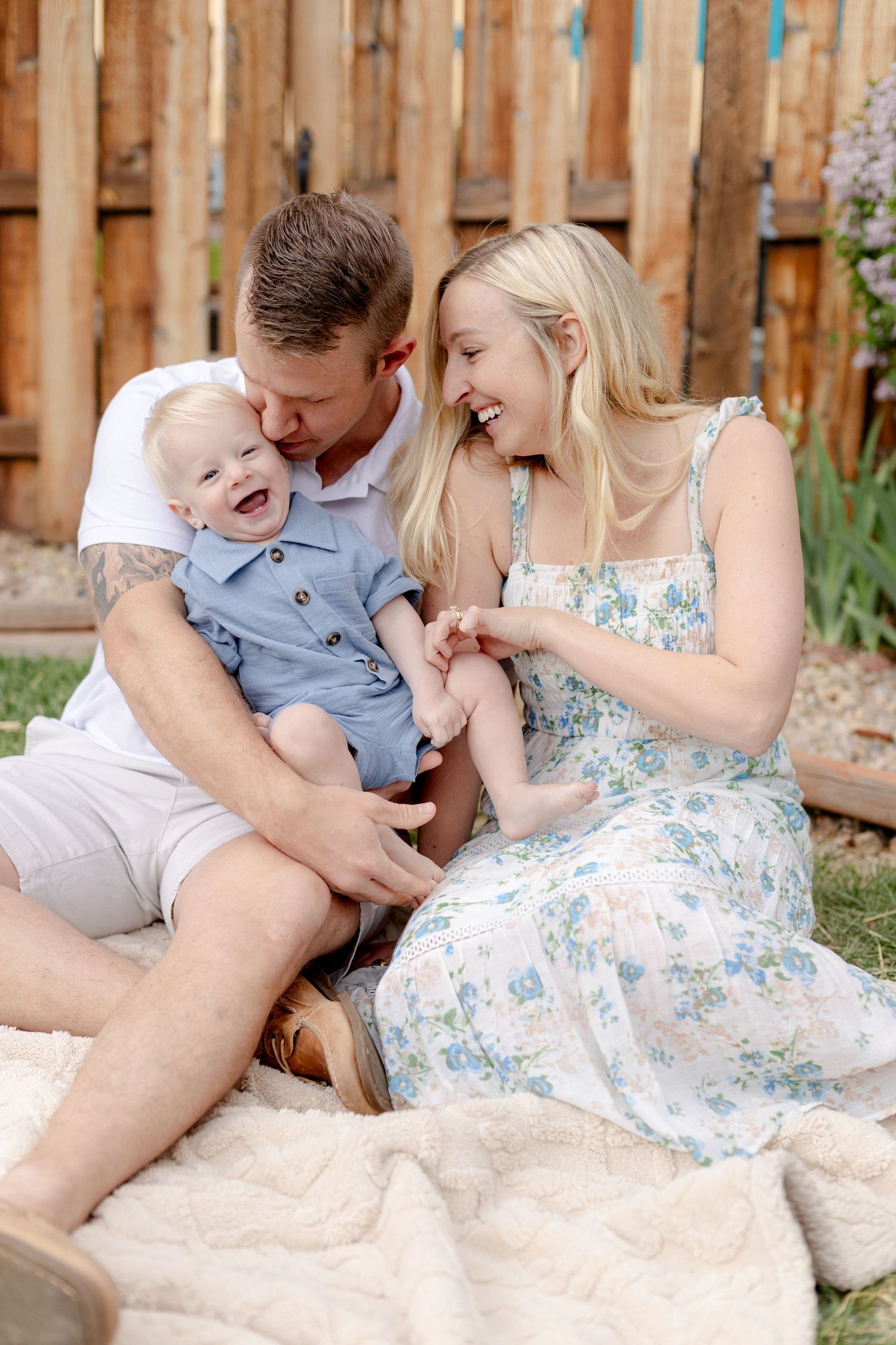 Happy family sitting on blanket in backyard, parents laughing with smiling baby boy in blue outfit on sunny day.
