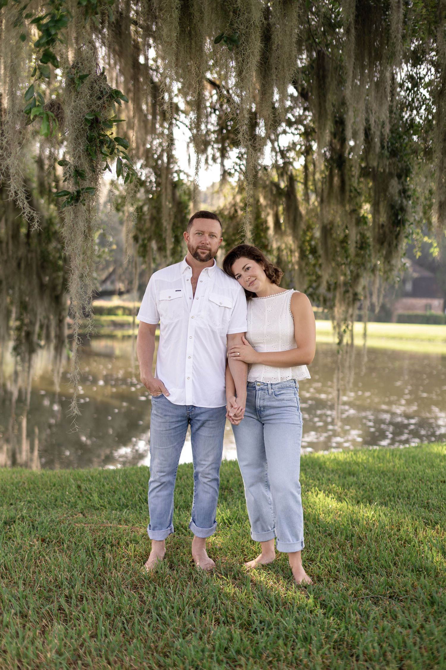 Couple posing barefoot by a pond under Spanish moss-draped trees on green grass.