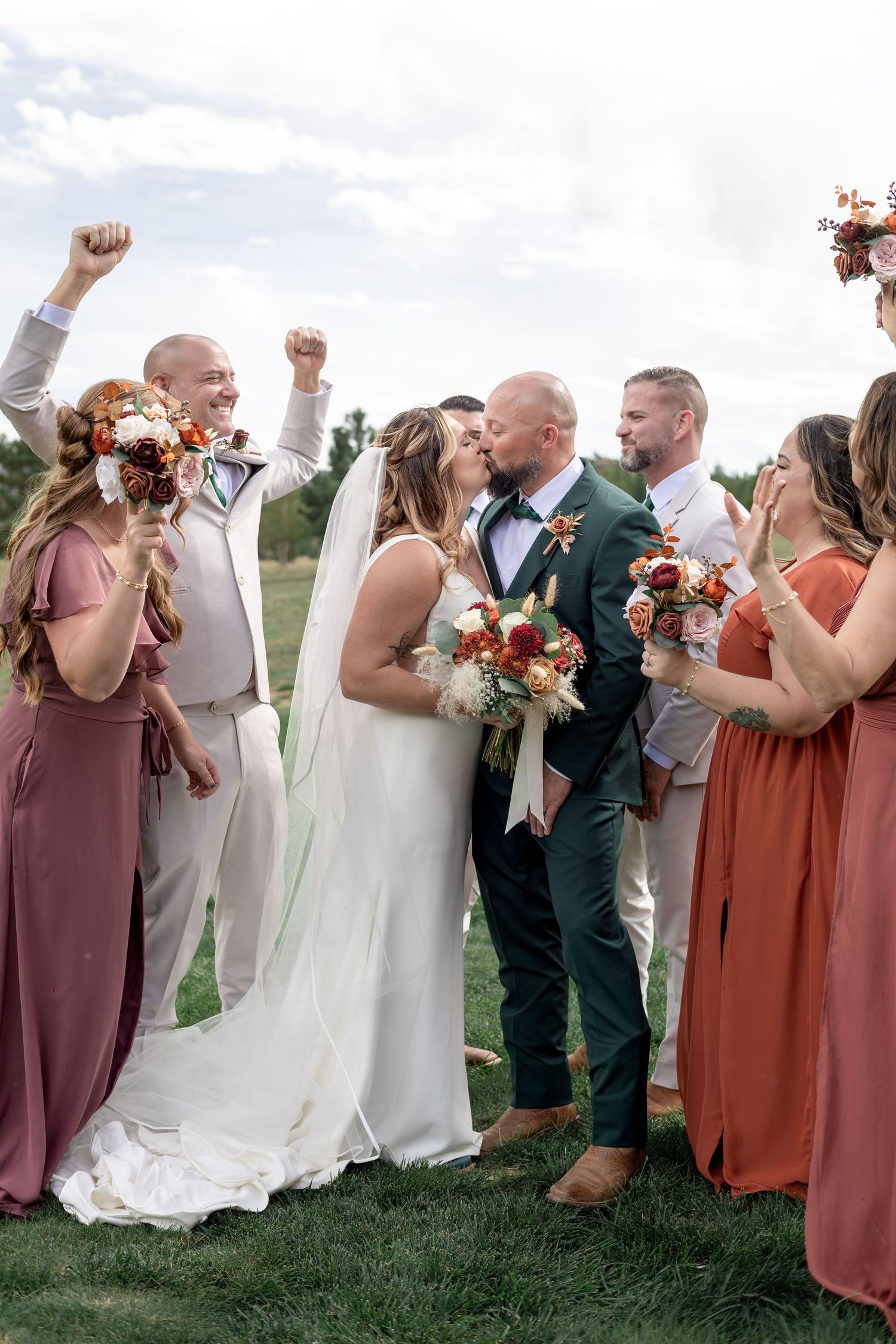 Bride and groom share a kiss surrounded by cheering wedding party in rust and mauve bridesmaid dresses outdoors.