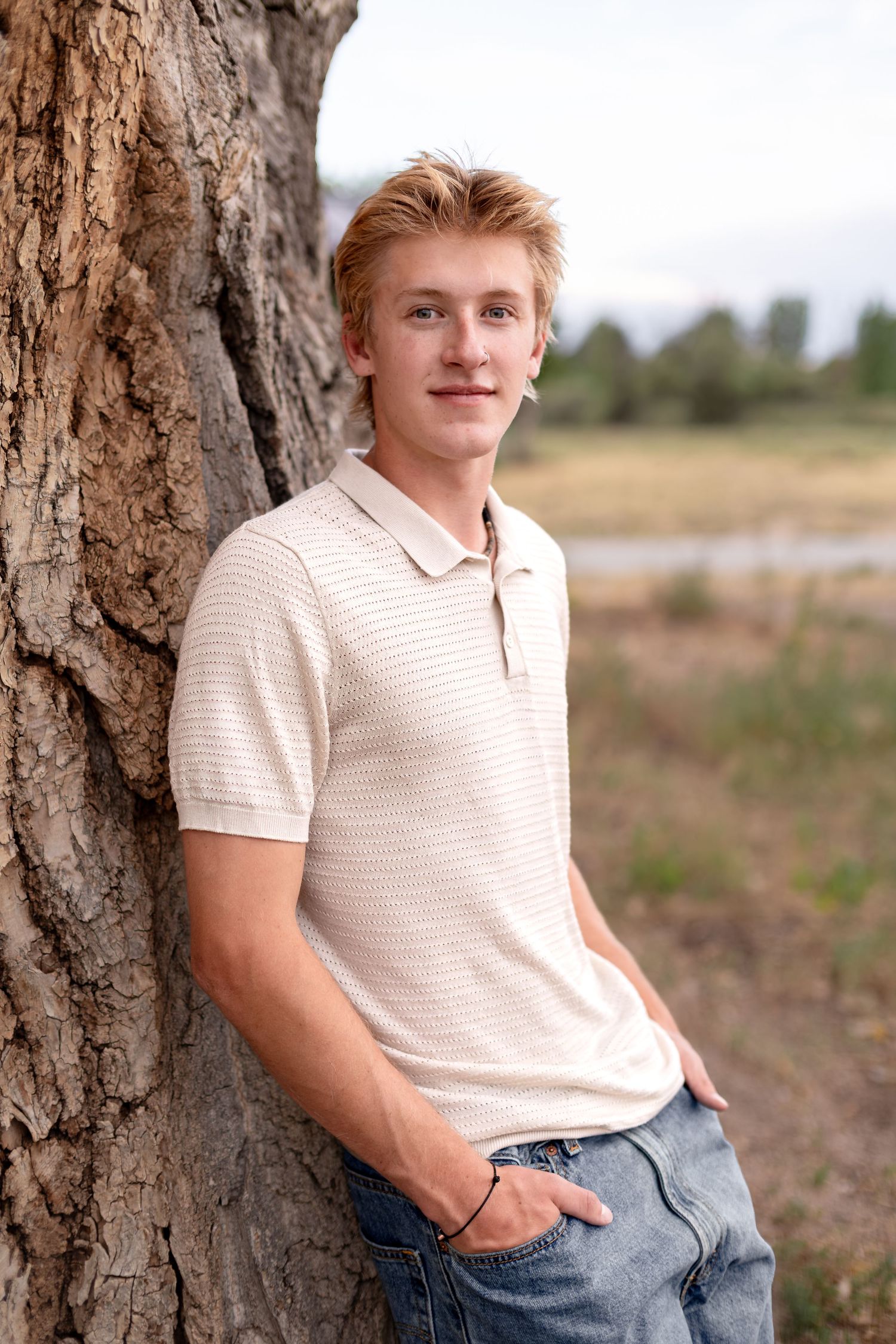 Young blond teen boy leaning against a large tree trunk in a field, wearing a polo shirt and jeans.