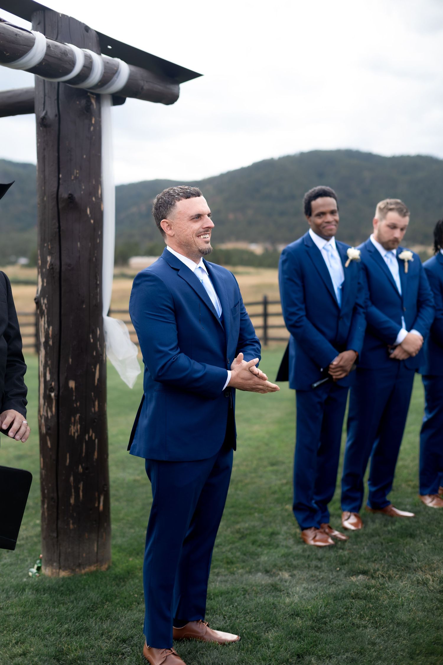 Groom in navy blue suit smiling at altar during outdoor wedding ceremony with groomsmen standing behind him.
