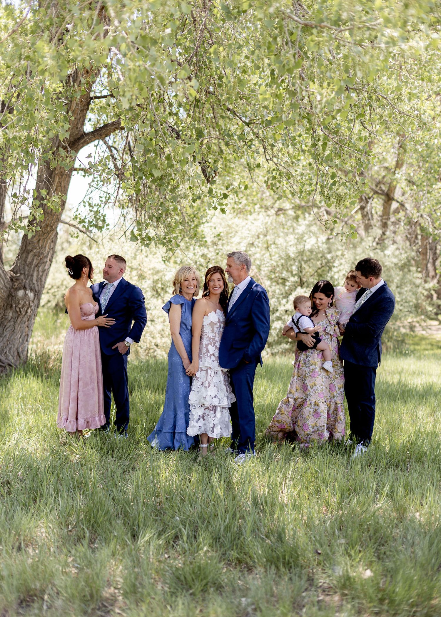 Family group portrait in elegant formal attire standing among green trees and grass at an outdoor wedding celebration.