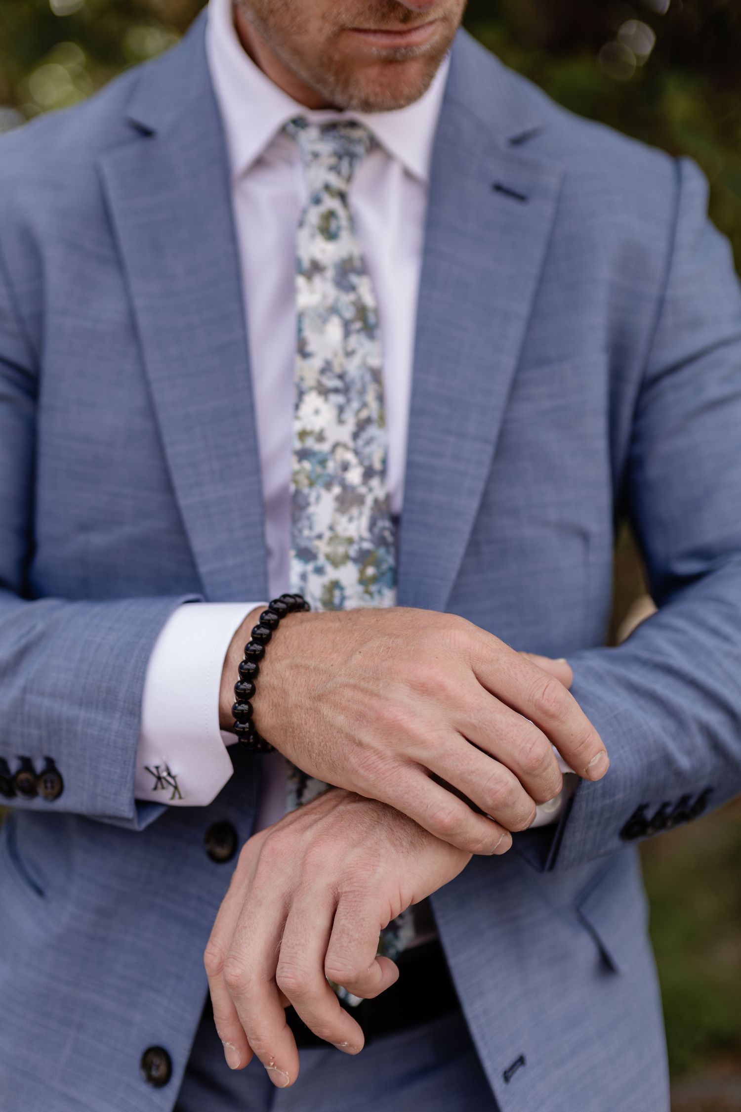 Groom in blue suit adjusting cuff with floral tie and black beaded bracelet outdoors.