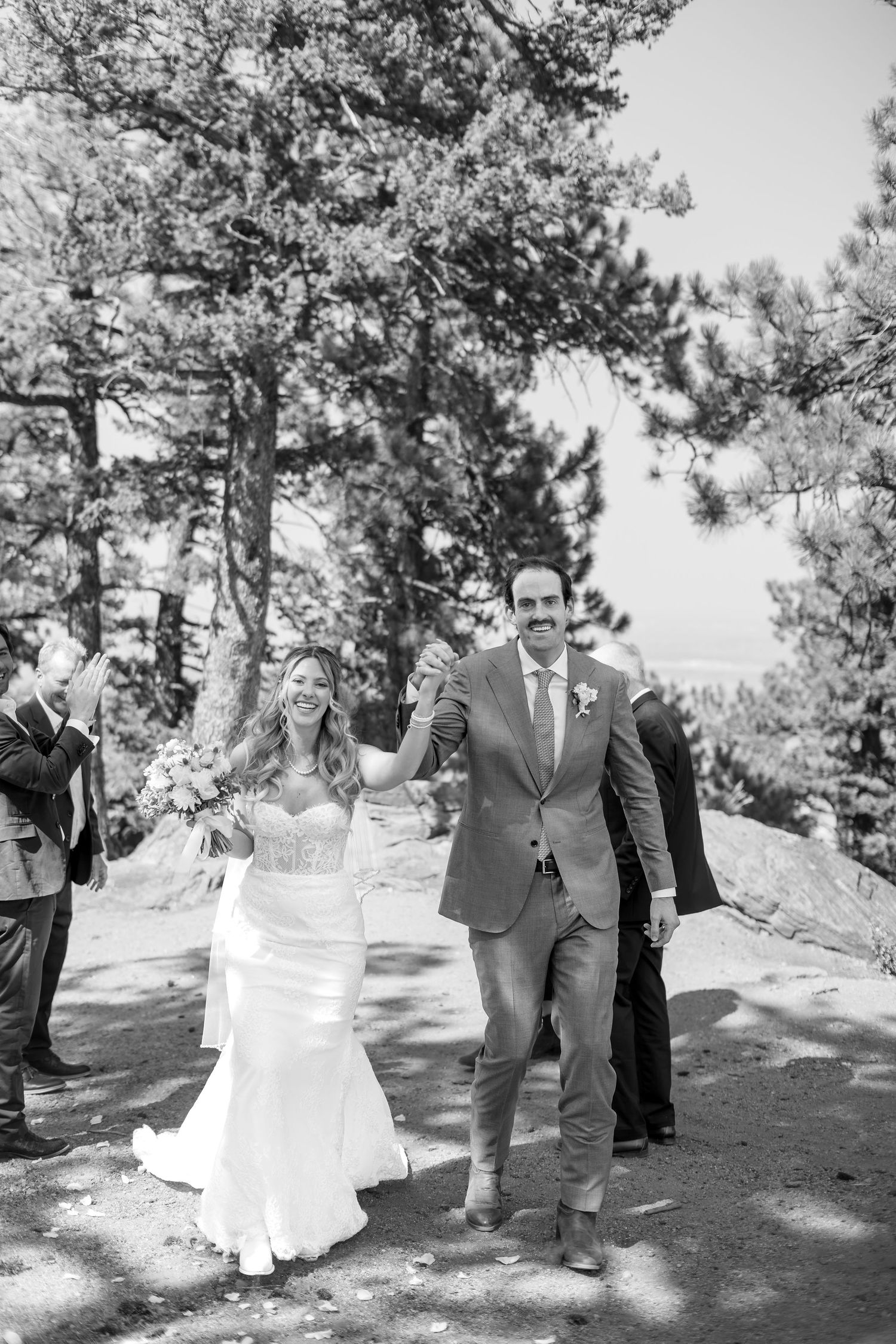 Black and white photo of a joyful bride and groom walking hand in hand after their outdoor forest wedding ceremony.