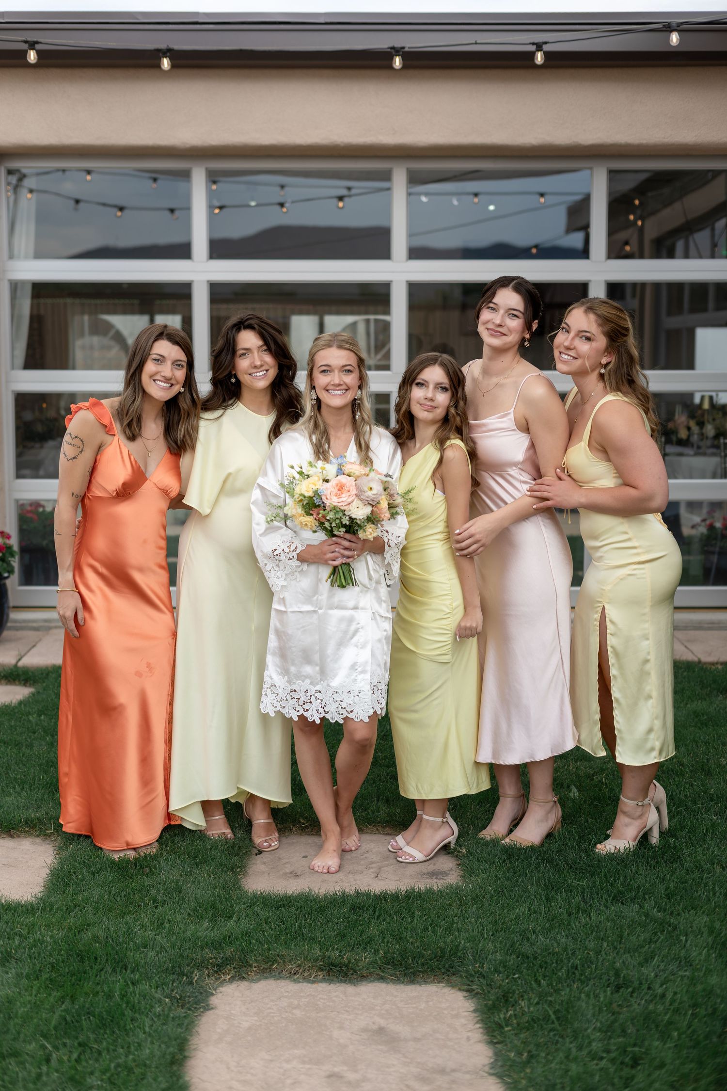Bride in white robe holds bouquet with six bridesmaids in colorful pastel gowns posing outdoors at wedding venue.