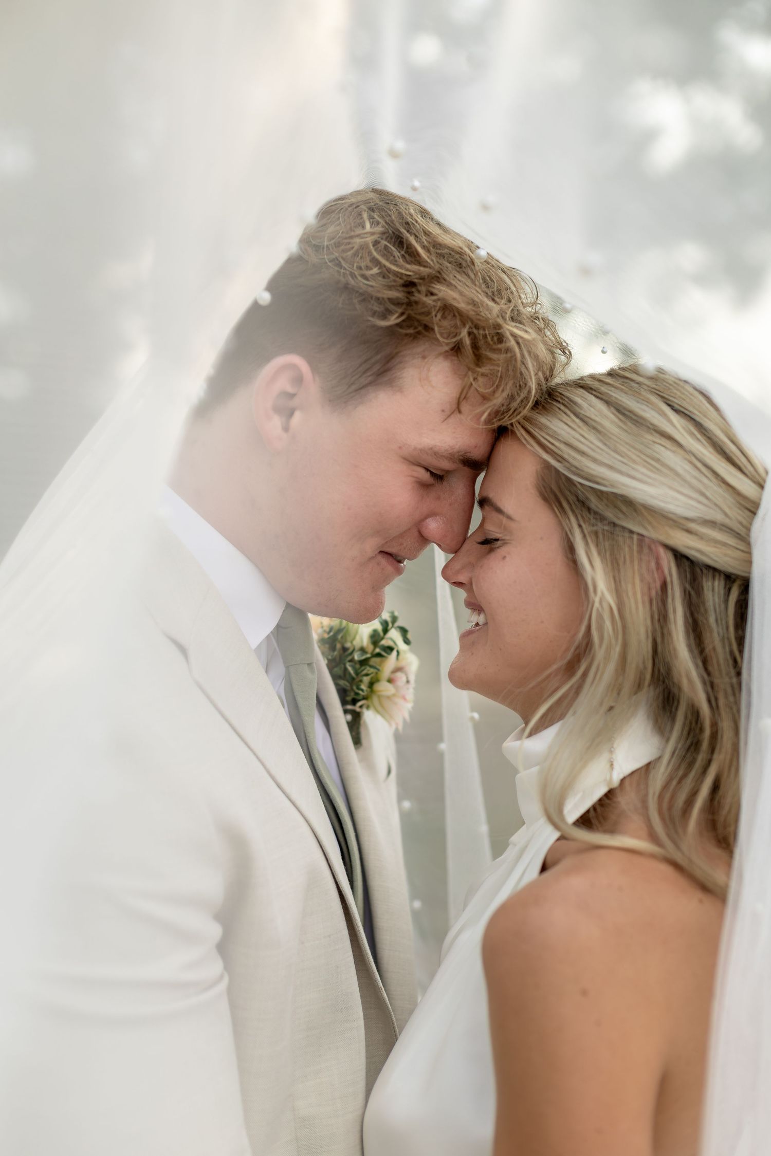 Bride and groom touching foreheads tenderly under a veil, sharing an intimate moment on their wedding day.