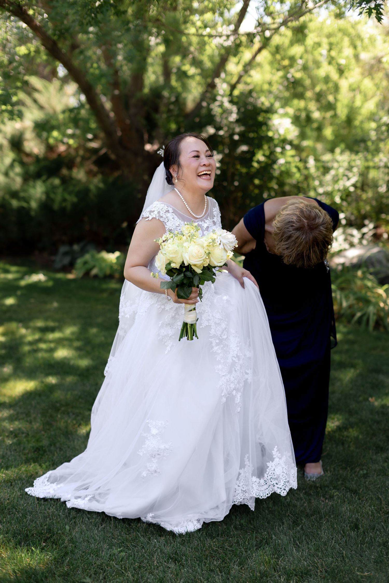 Smiling bride in white lace gown holds yellow bouquet while woman in blue dress adjusts her wedding dress outdoors.