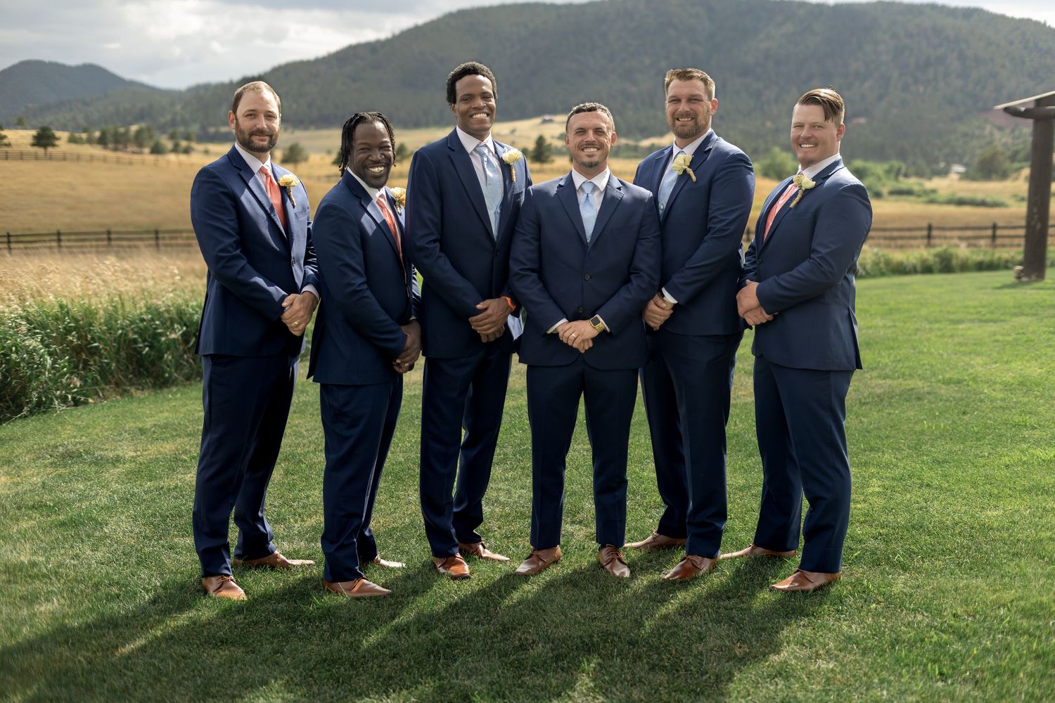 Six groomsmen in navy suits with coral ties pose outdoors at a mountain wedding venue on a sunny day.