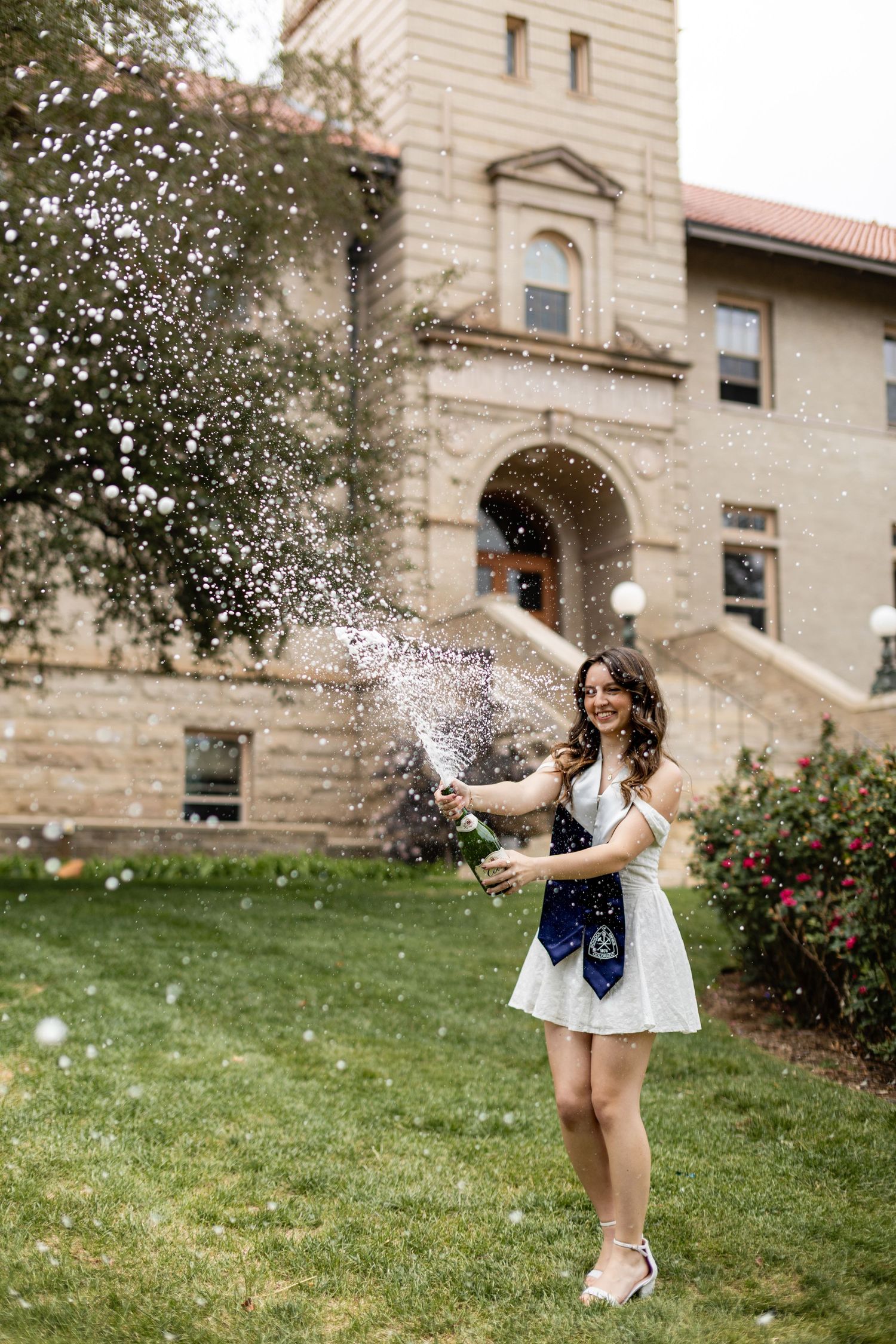 A joyful graduate in a white dress pops champagne outside a university building, celebrating her achievement.
