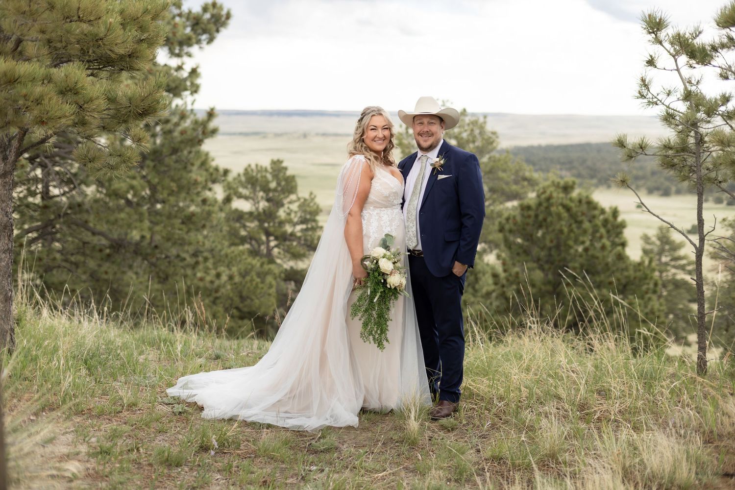Bride and groom smiling outdoors on a scenic hillside, groom wearing a cowboy hat and navy suit.