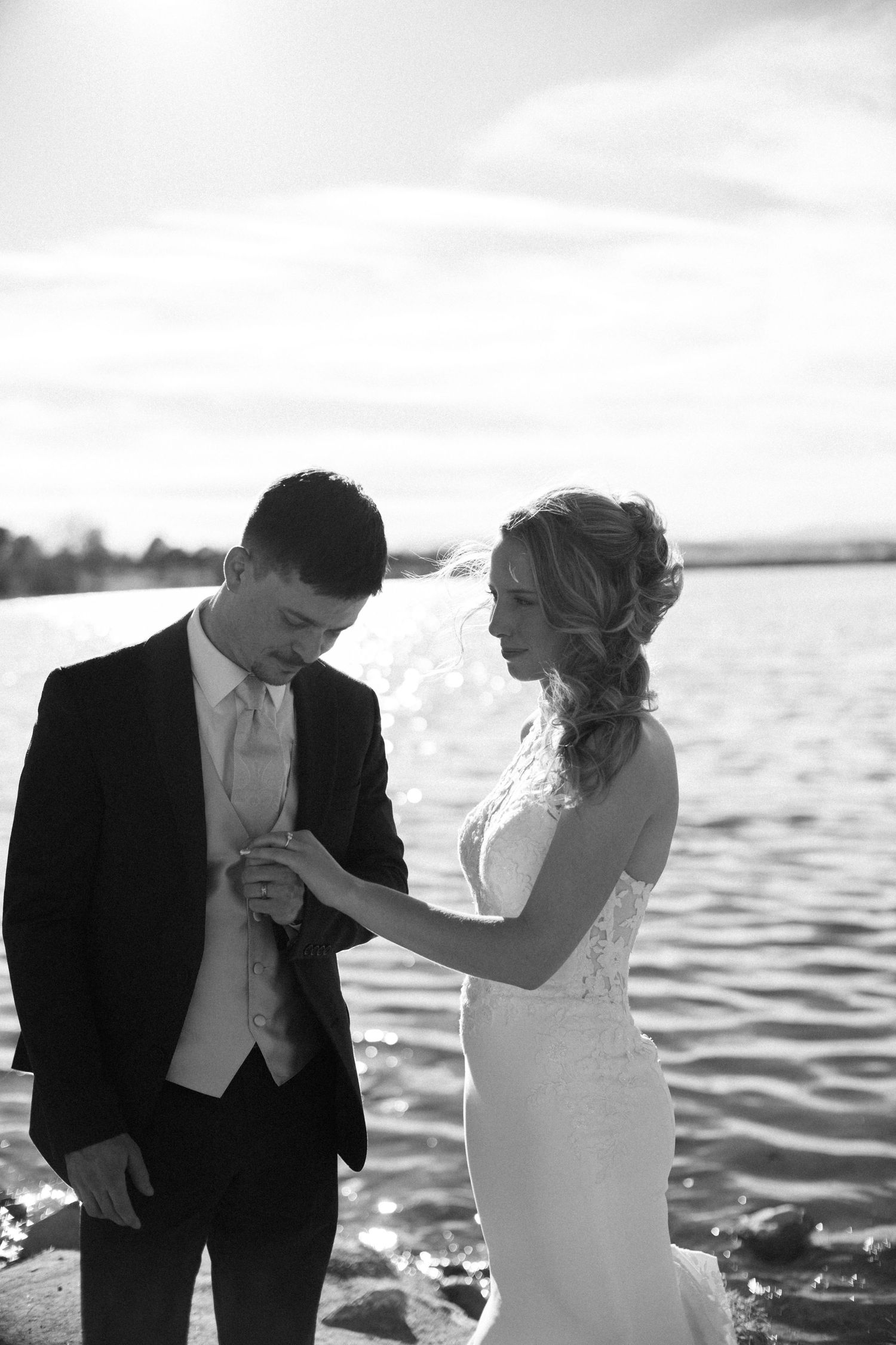 Black and white photo of bride placing wedding ring on grooms finger beside a sunlit lake.
