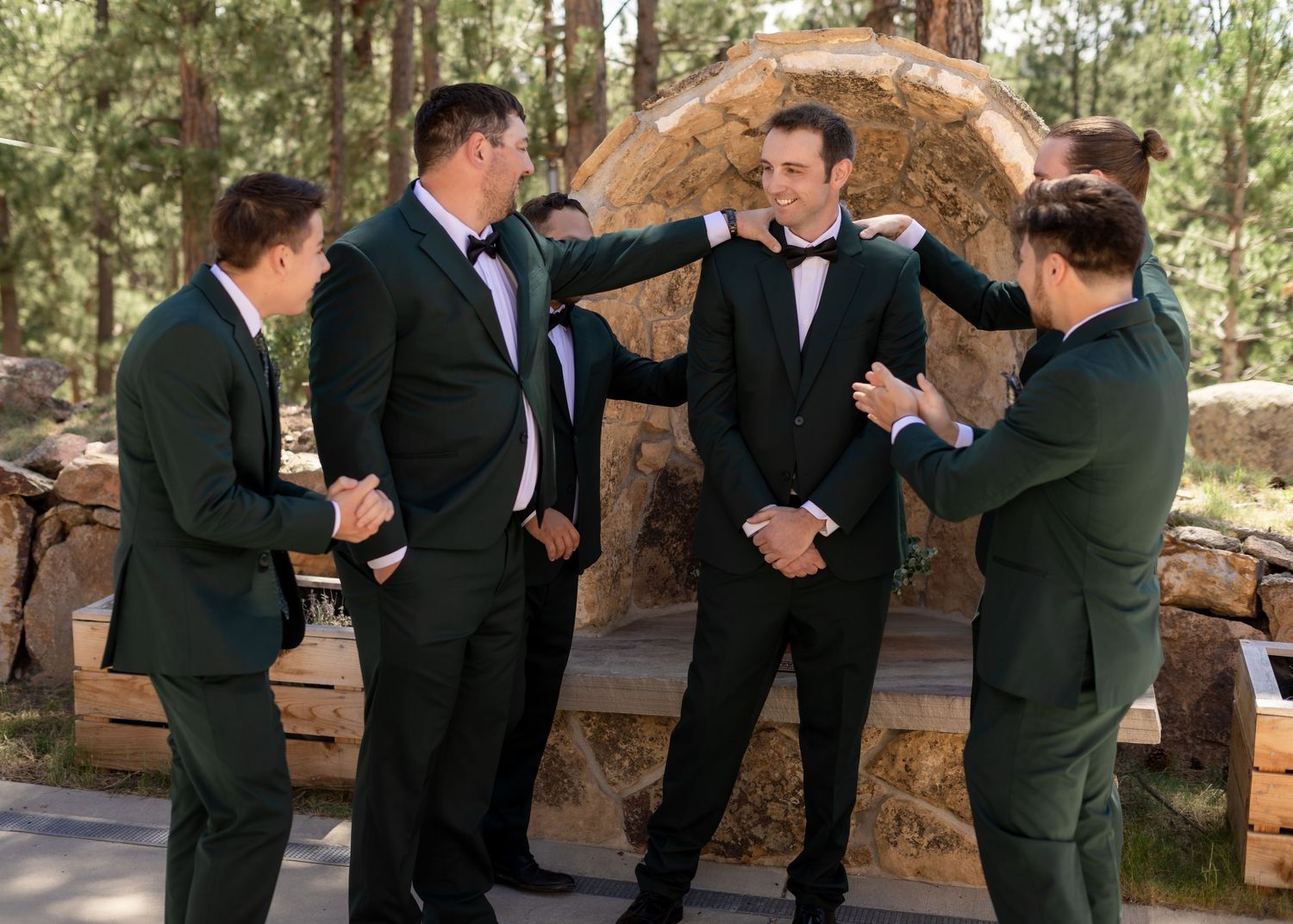 Groom in black tuxedo celebrates with groomsmen in dark green suits outdoors at a rustic woodland wedding venue.