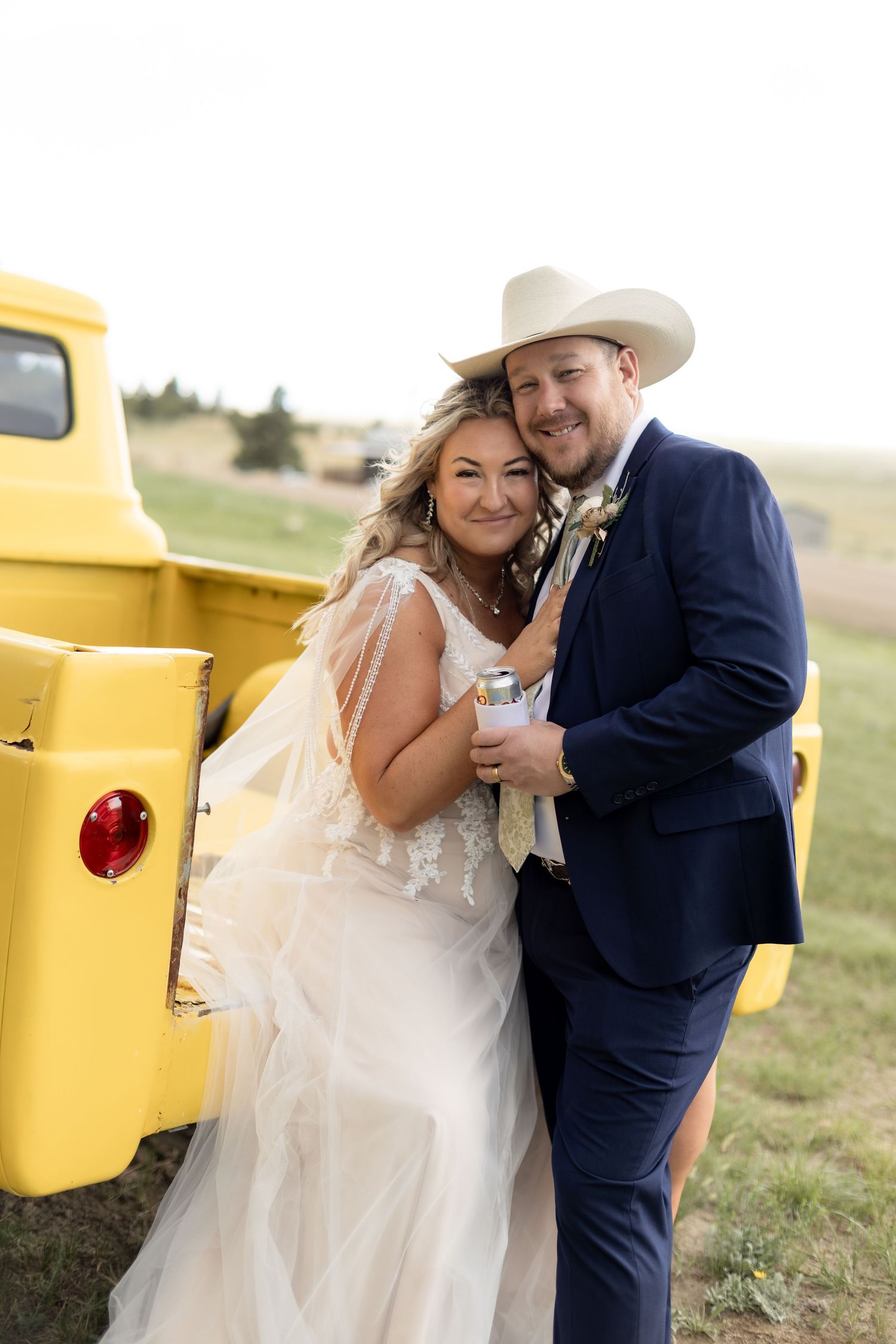 Smiling bride and groom pose together beside a vintage yellow truck at their country wedding.