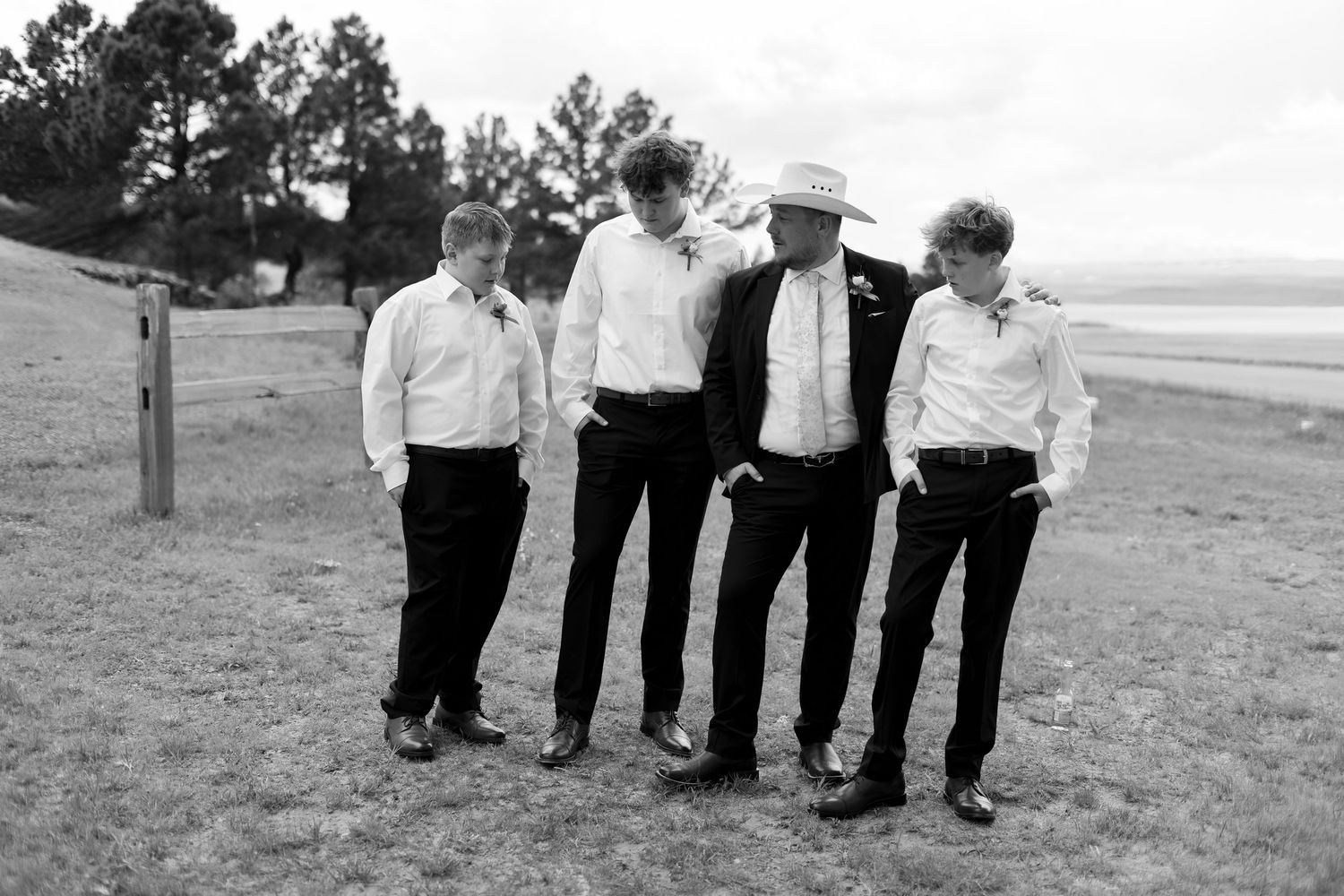 Four groomsmen in black suits and white shirts walk together outdoors at a rustic wedding venue.