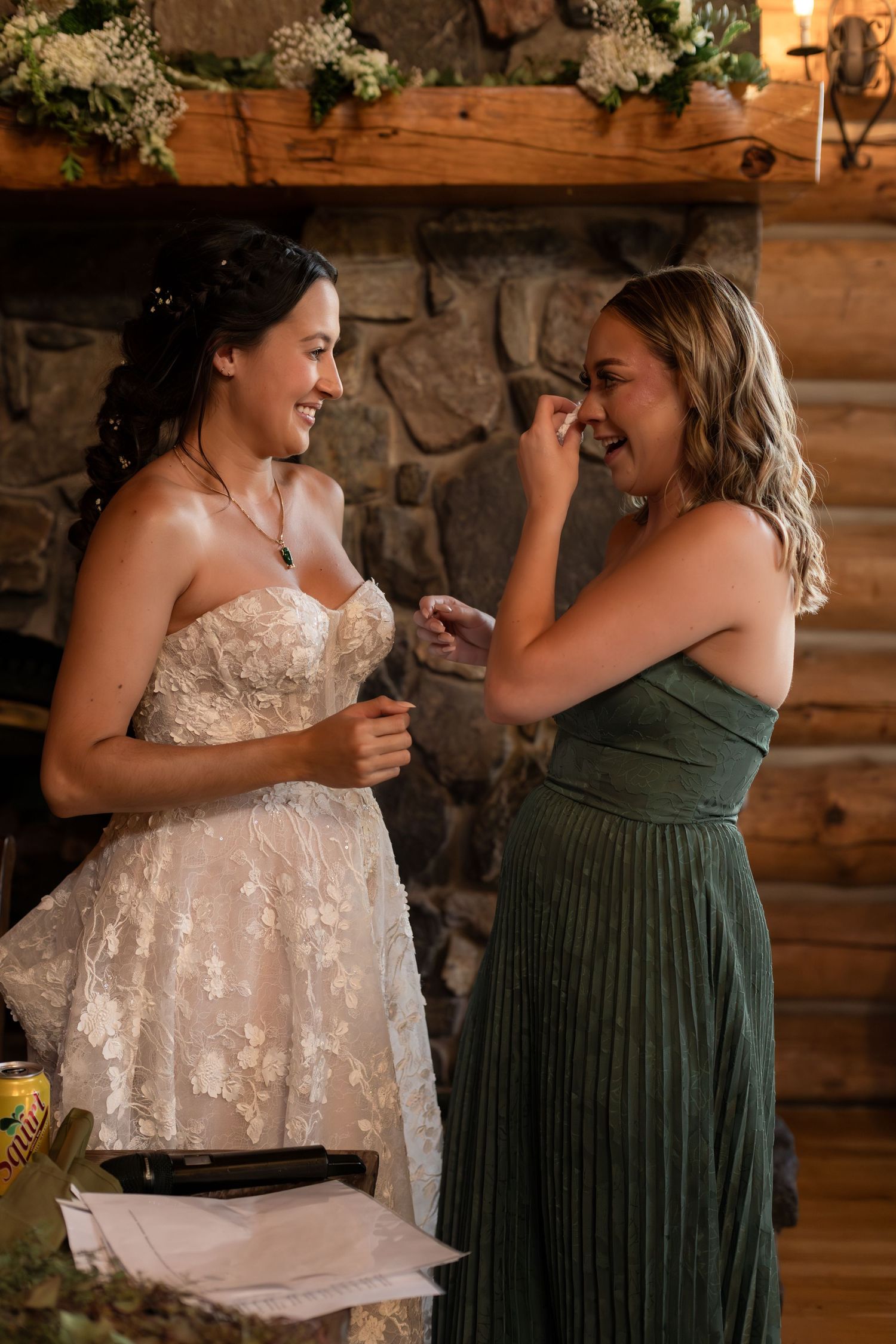 Bride in lace gown laughing with bridesmaid in green dress near stone fireplace at rustic wedding venue.