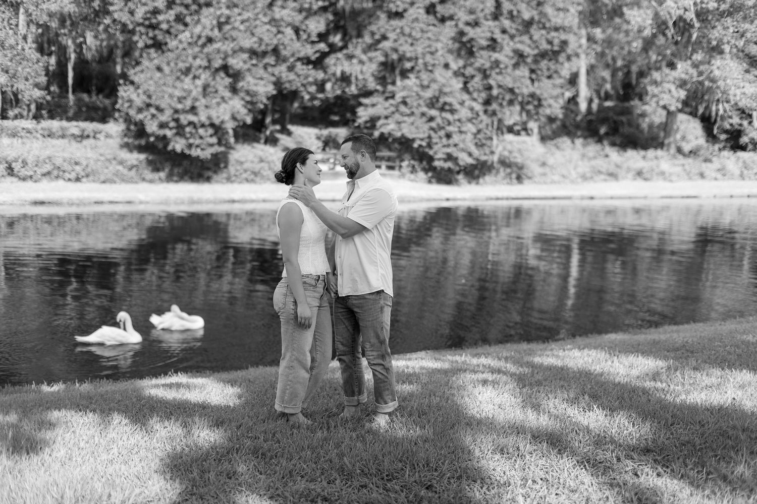 Black and white photo of a couple embracing by a serene pond with swans, surrounded by lush trees.