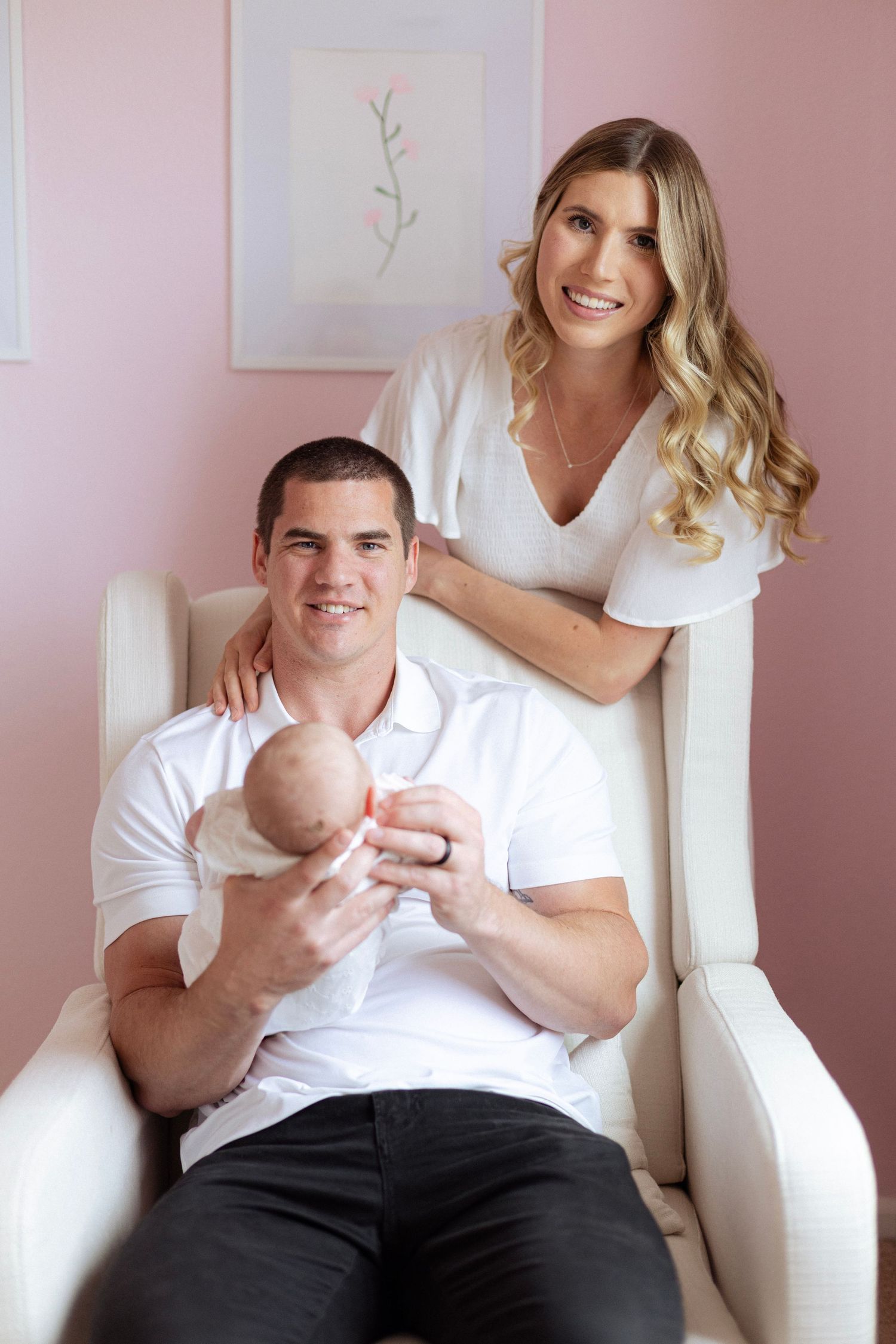 Happy young parents in white outfits sitting in a nursery chair, lovingly holding their newborn baby.