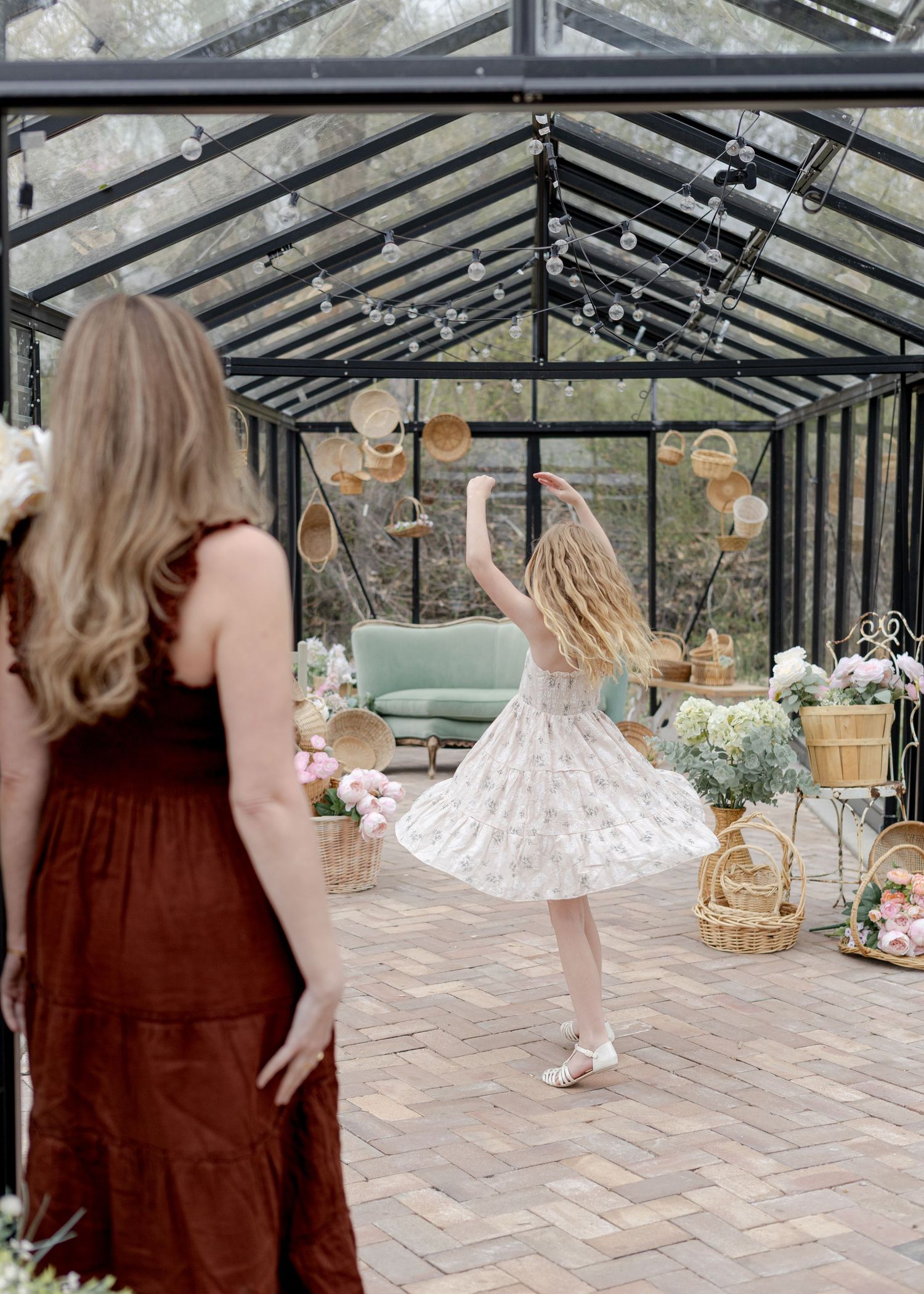 A girl in a floral dress twirls in a glass greenhouse while a woman in a rust dress watches nearby.