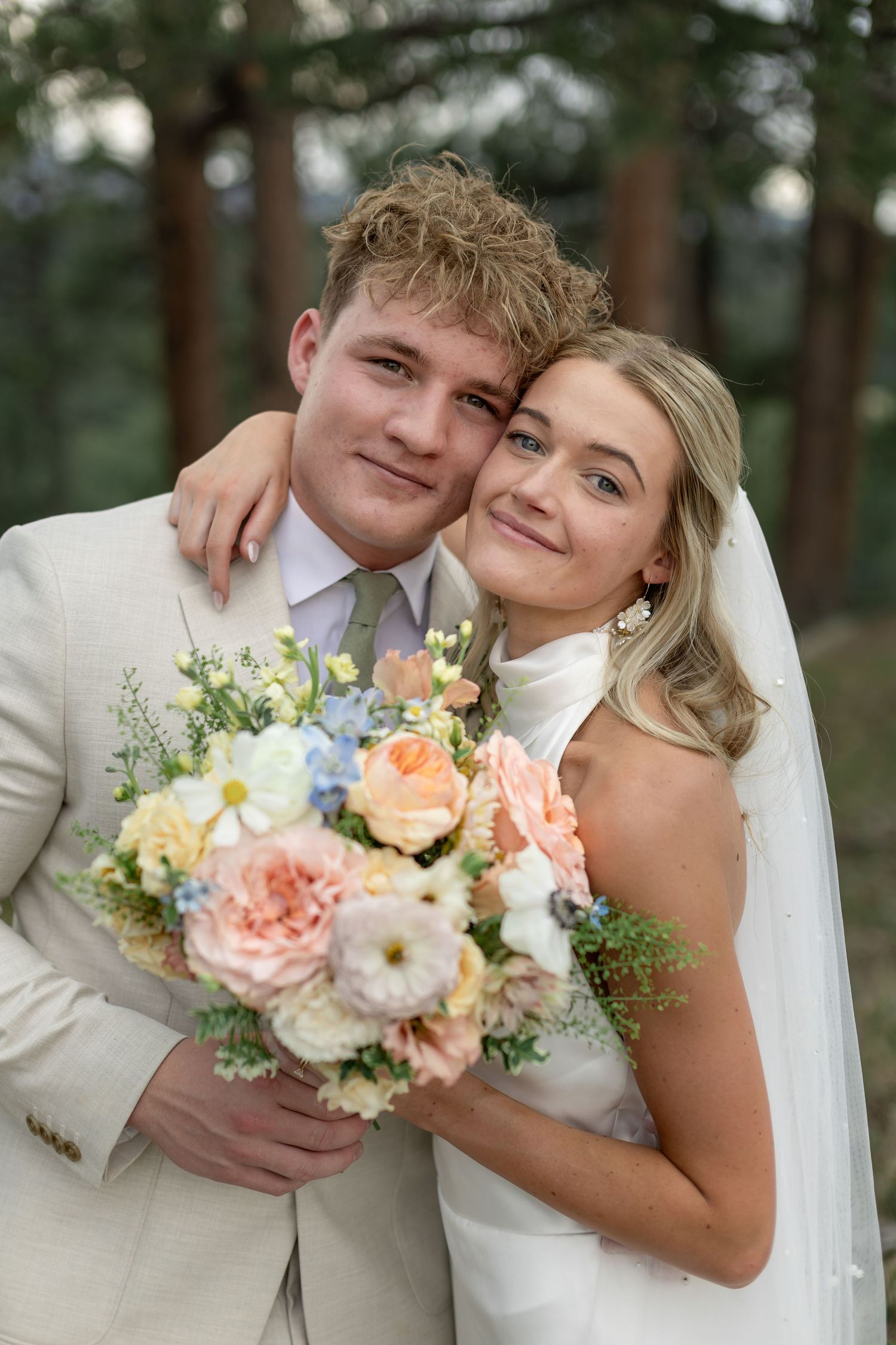 Smiling bride and groom posing together holding a pastel bouquet of pink, peach, and blue flowers outdoors.