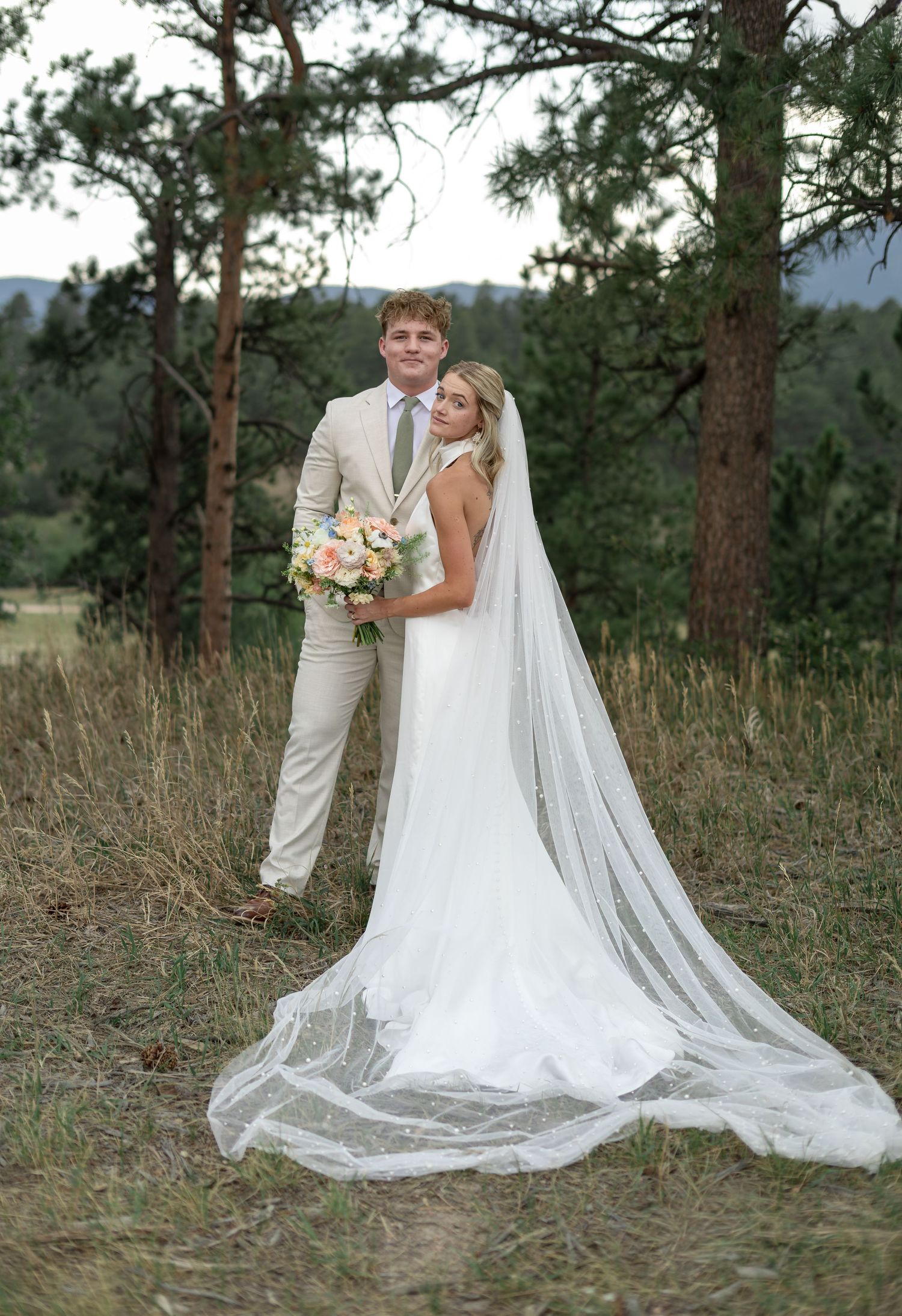 Bride in white gown with cathedral veil holds pastel bouquet while groom stands behind her outdoors.