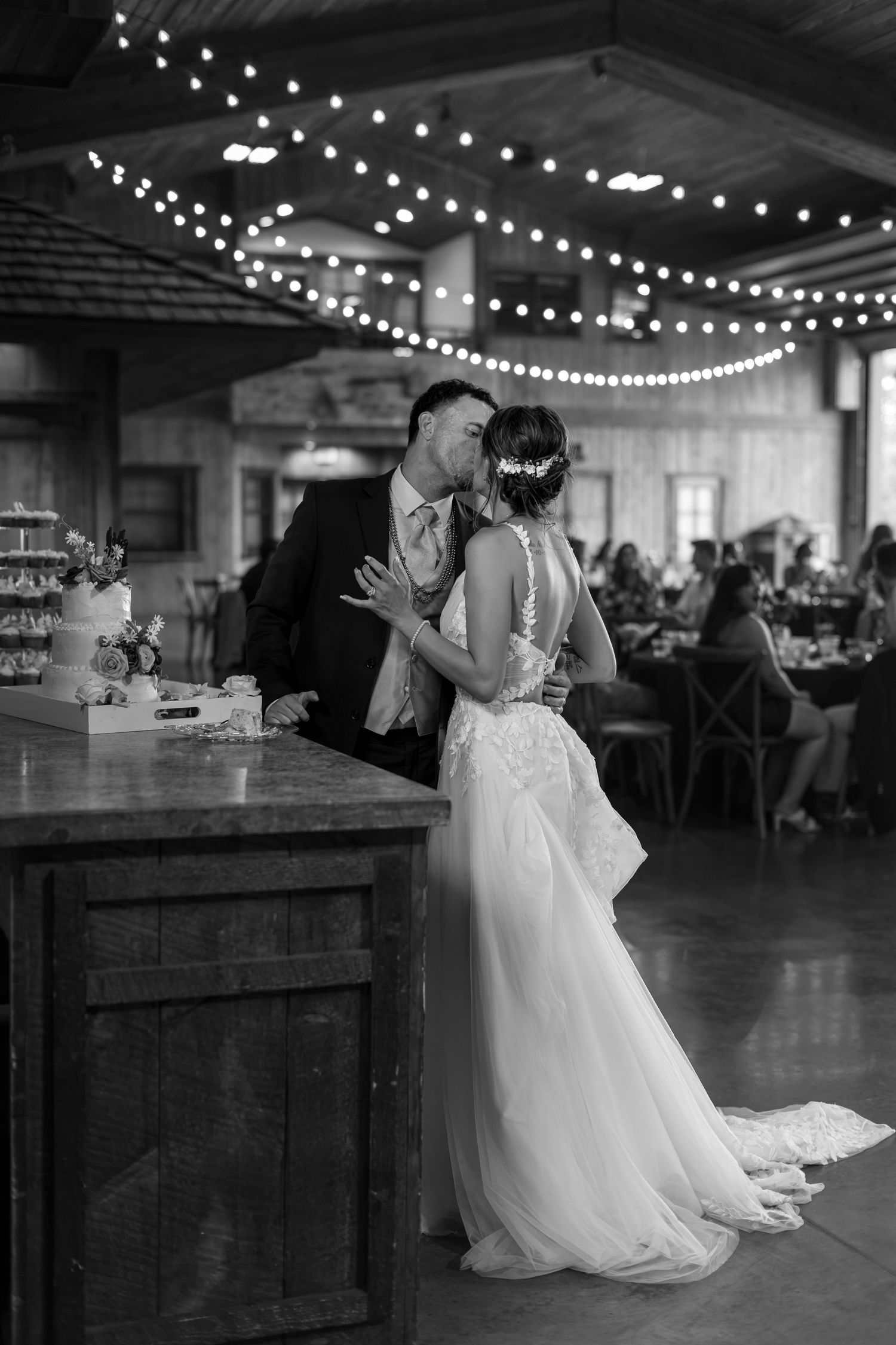 Black and white photo of bride and groom kissing near wedding cake under string lights.