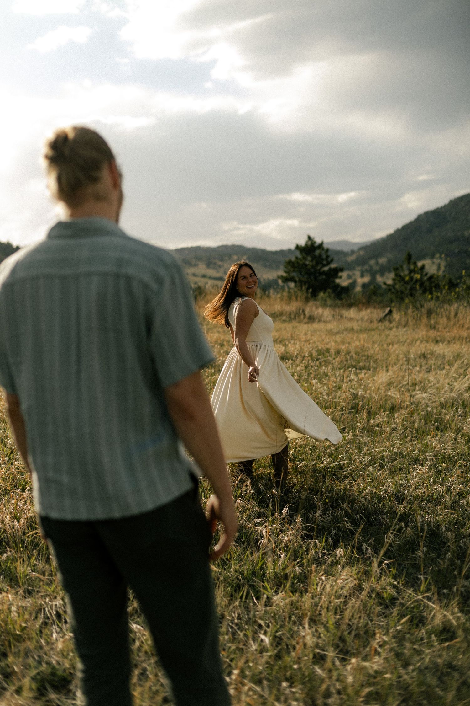 Woman in cream dress twirling in meadow while man watches, mountain backdrop.