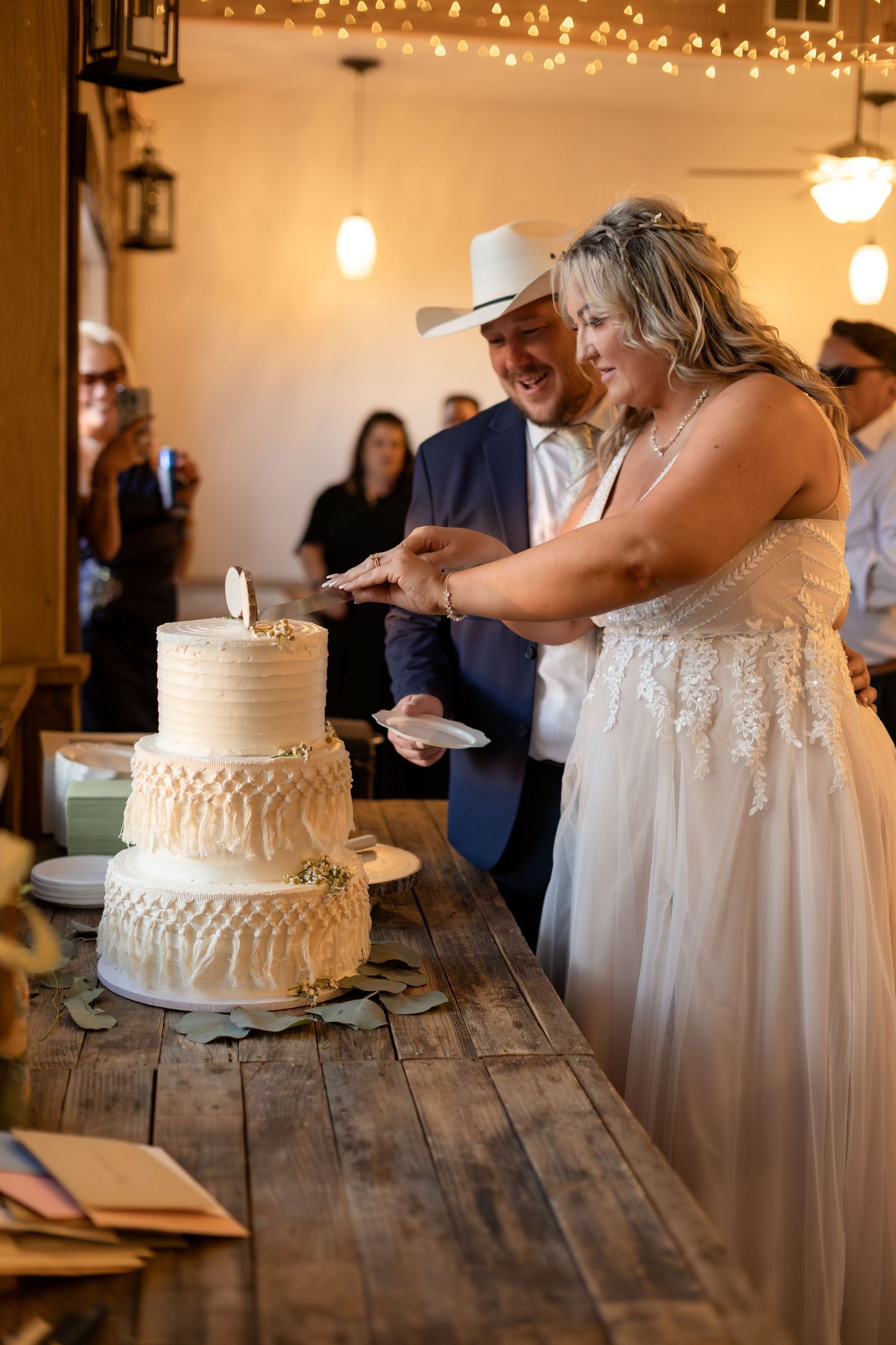 Bride and groom in cowboy hat cut three-tier white wedding cake at rustic barn reception.
