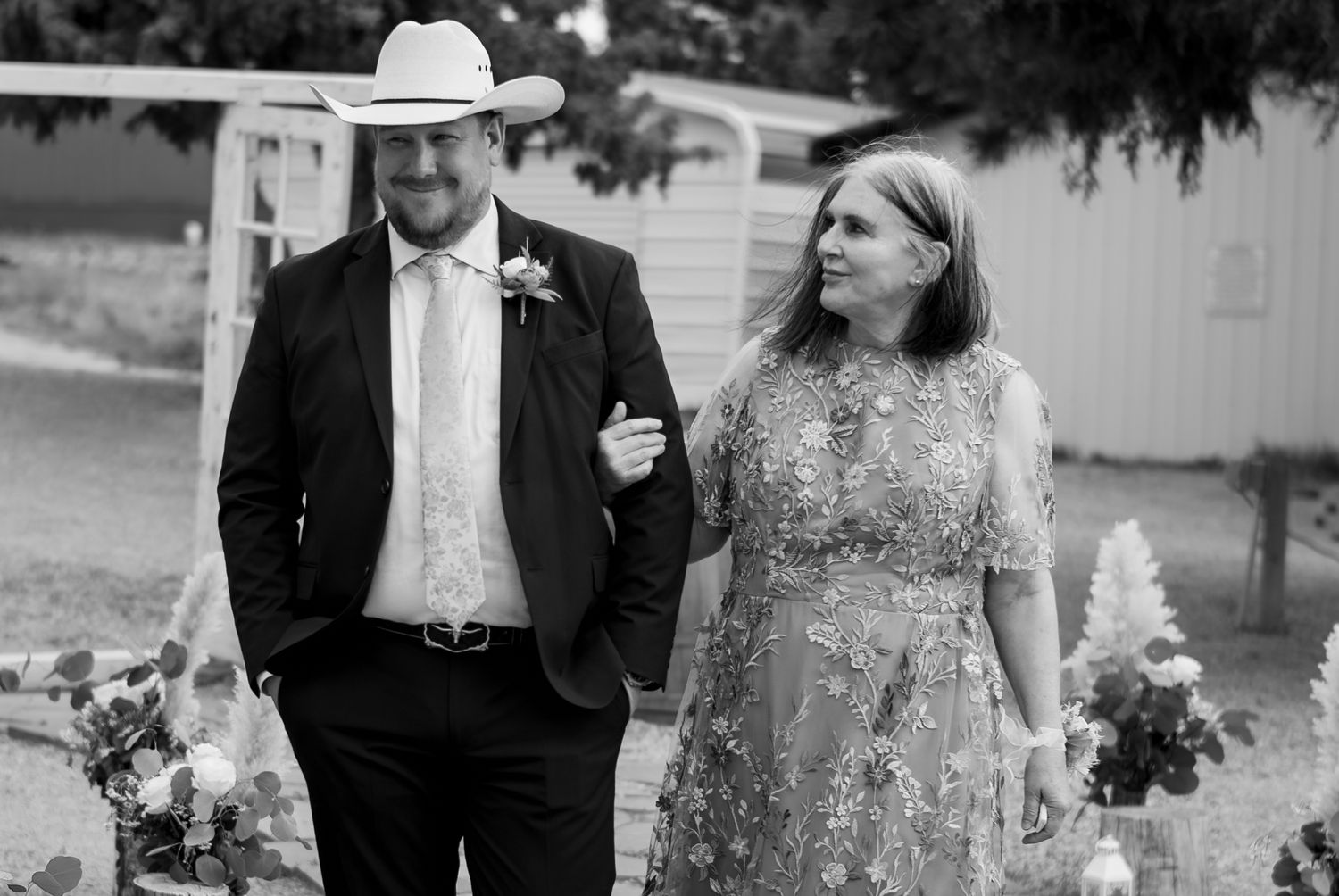 Black and white photo of man in cowboy hat and suit escorting woman in floral dress at wedding.
