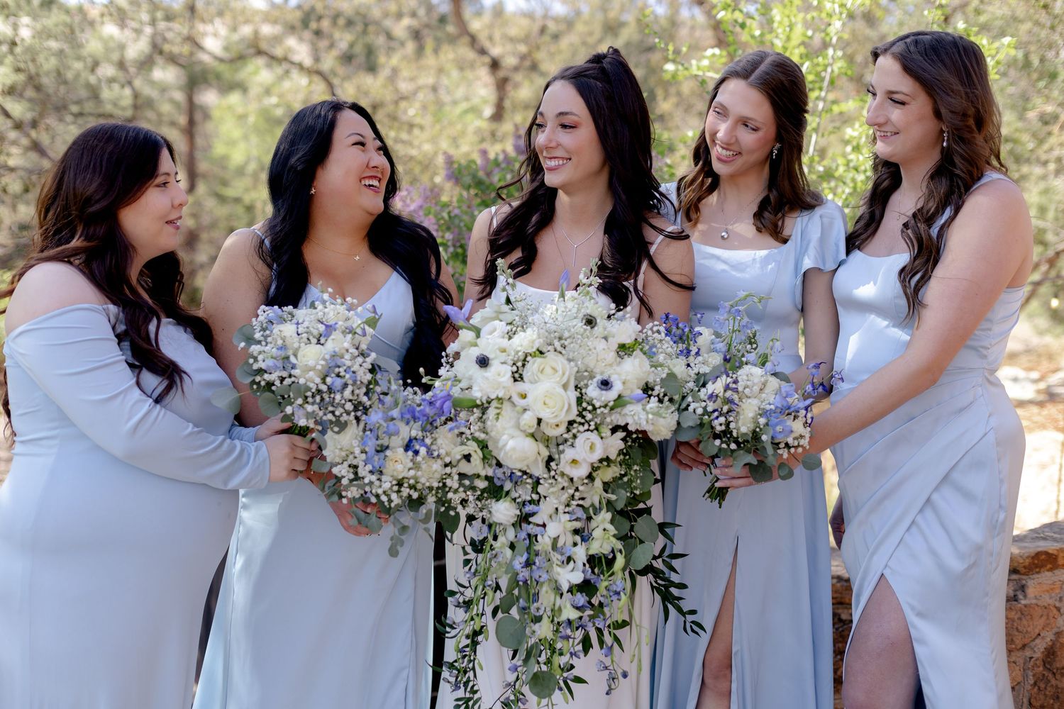 Five bridesmaids in light blue gowns laughing and holding white and blue floral bouquets outdoors at a wedding.
