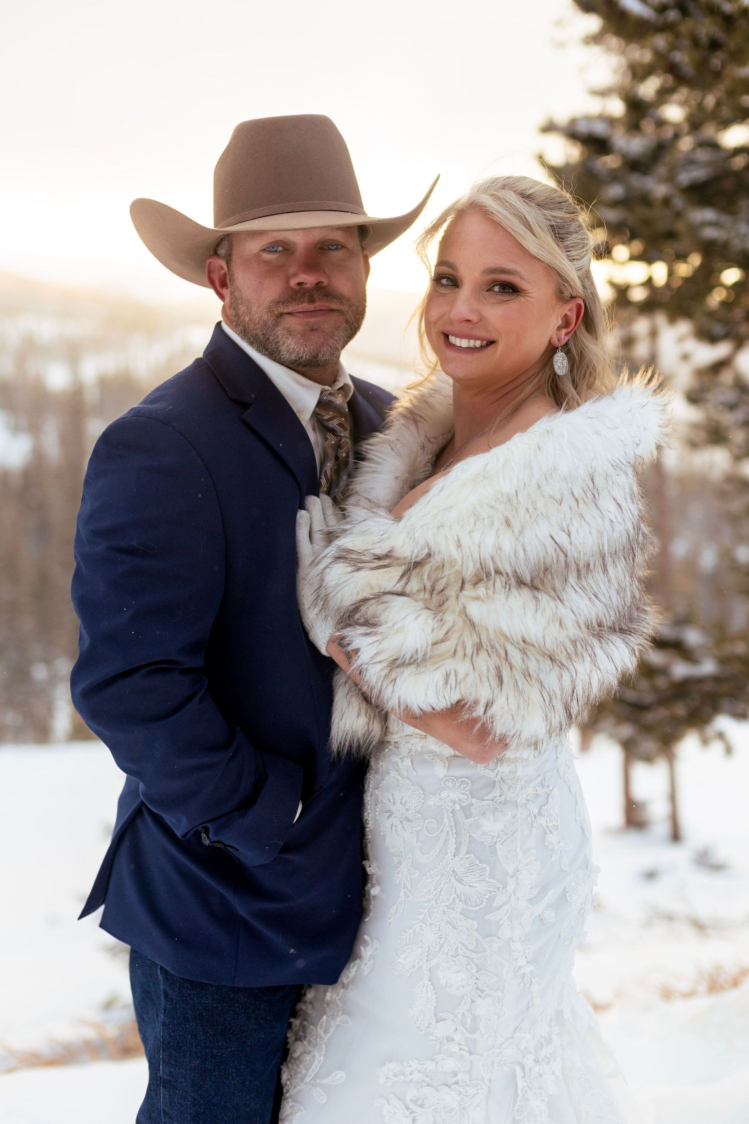 A winter wedding photo with the couple posing outdoors in the snow, with bride in white dress and groom in navy suit and cowboy hat.