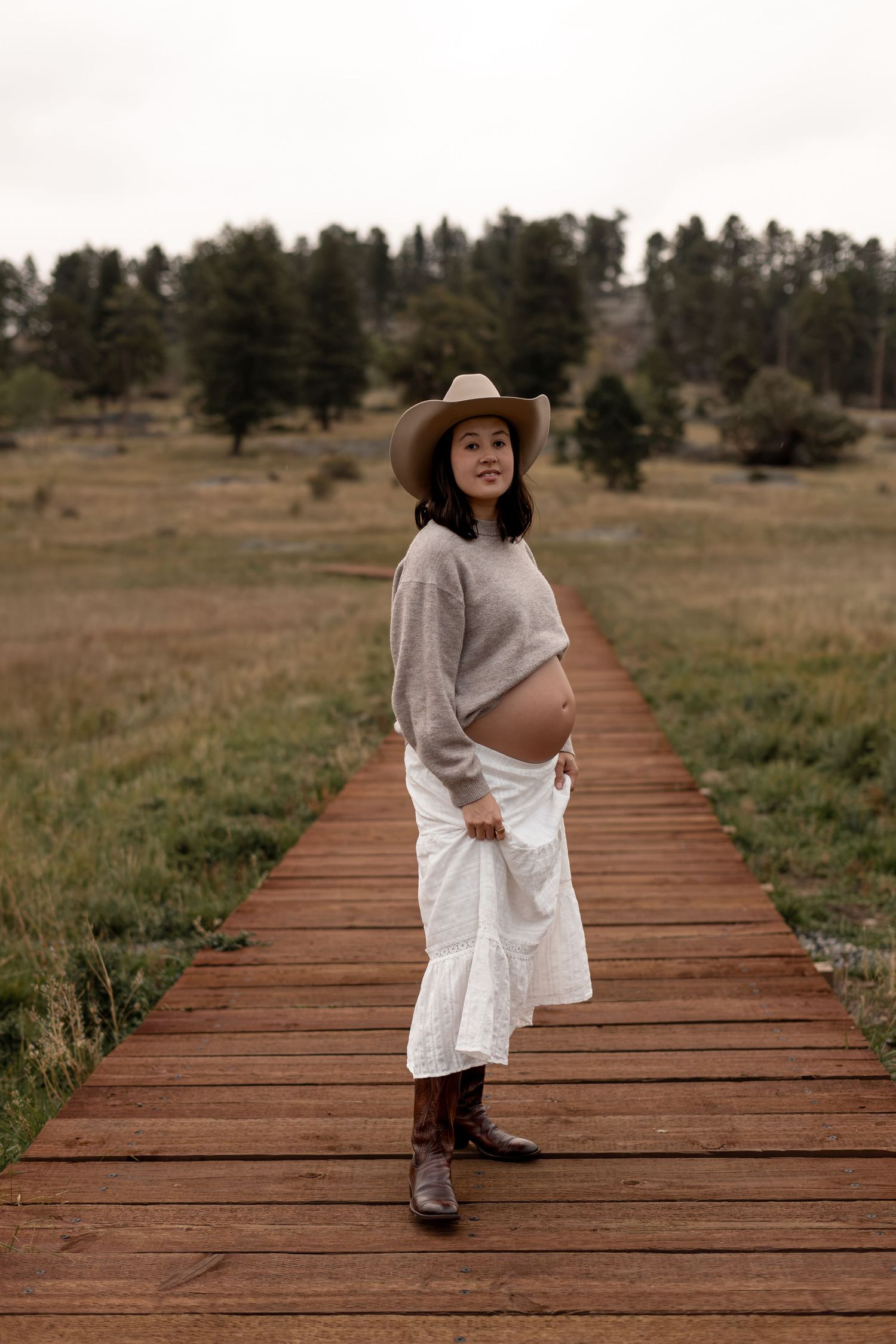 Pregnant woman in cowboy hat, gray sweater, white skirt, and boots stands on wooden boardwalk in rural meadow.