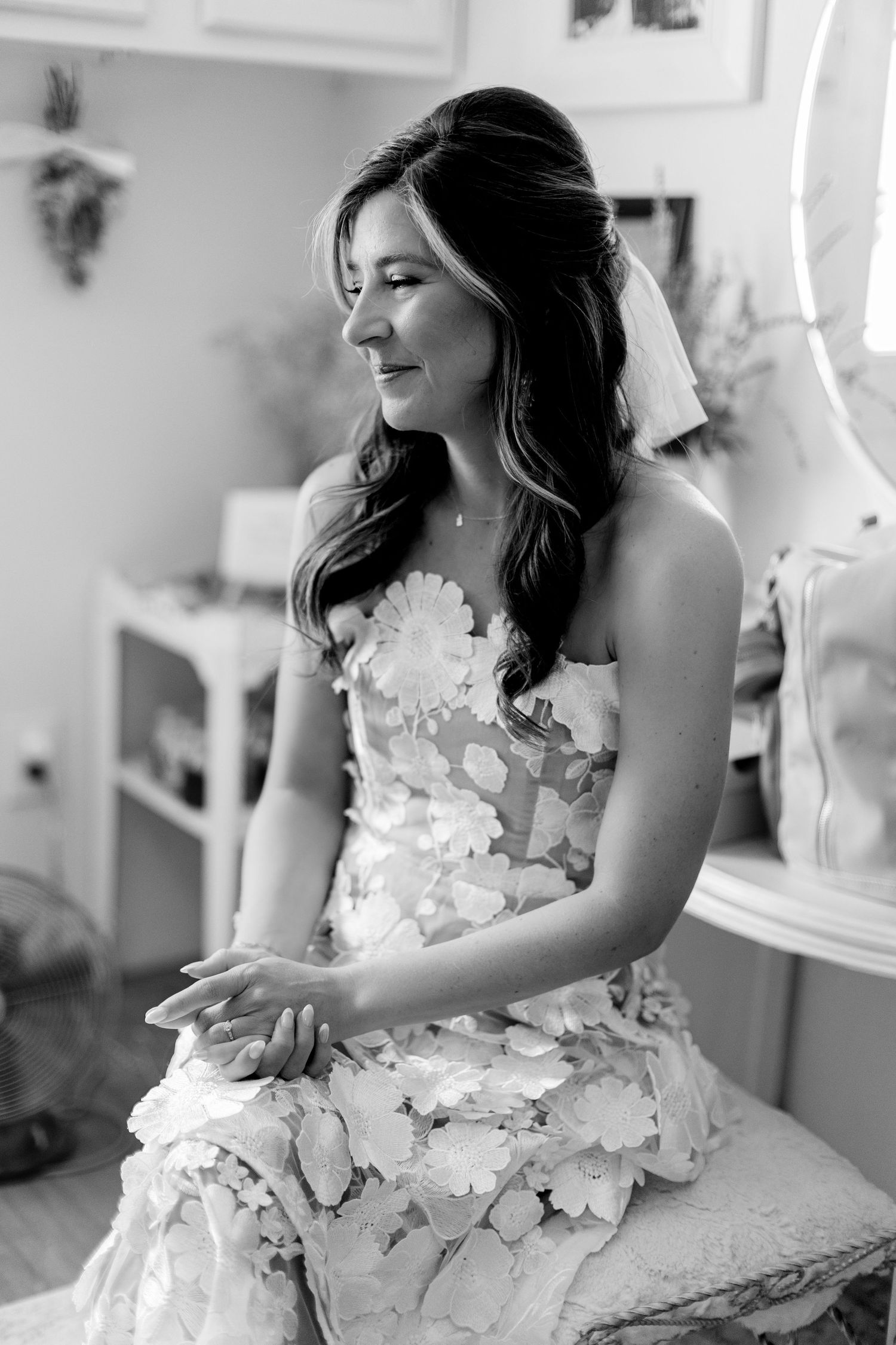 Elegant bride in floral applique gown sits and smiles softly in black and white portrait.