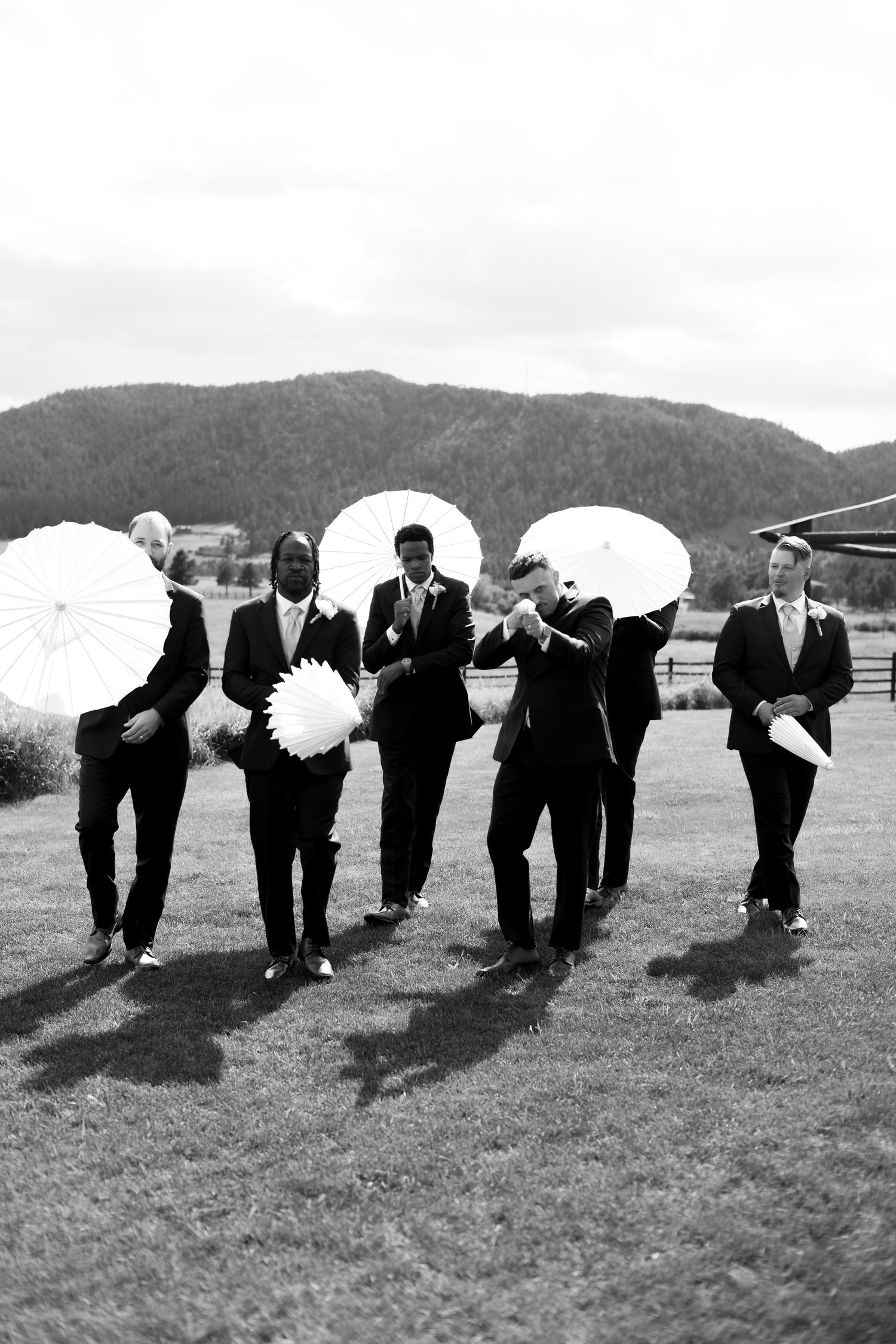 Groomsmen in black suits holding white parasols walk outdoors with mountain backdrop in black and white wedding photo.
