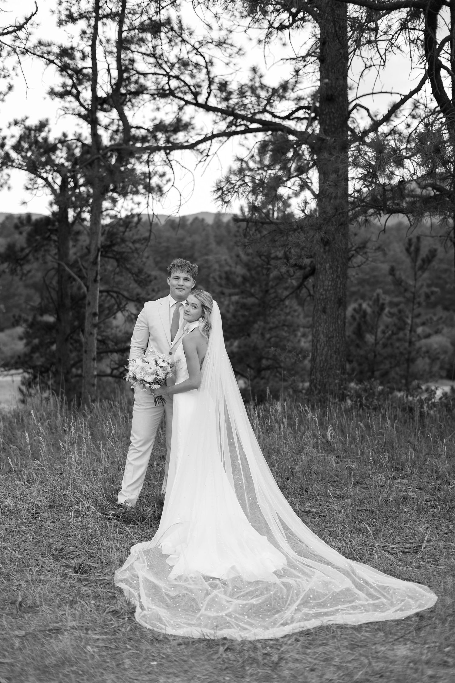 Black and white photo of bride and groom embracing outdoors among pine trees, bride wearing long veil trailing on grass.