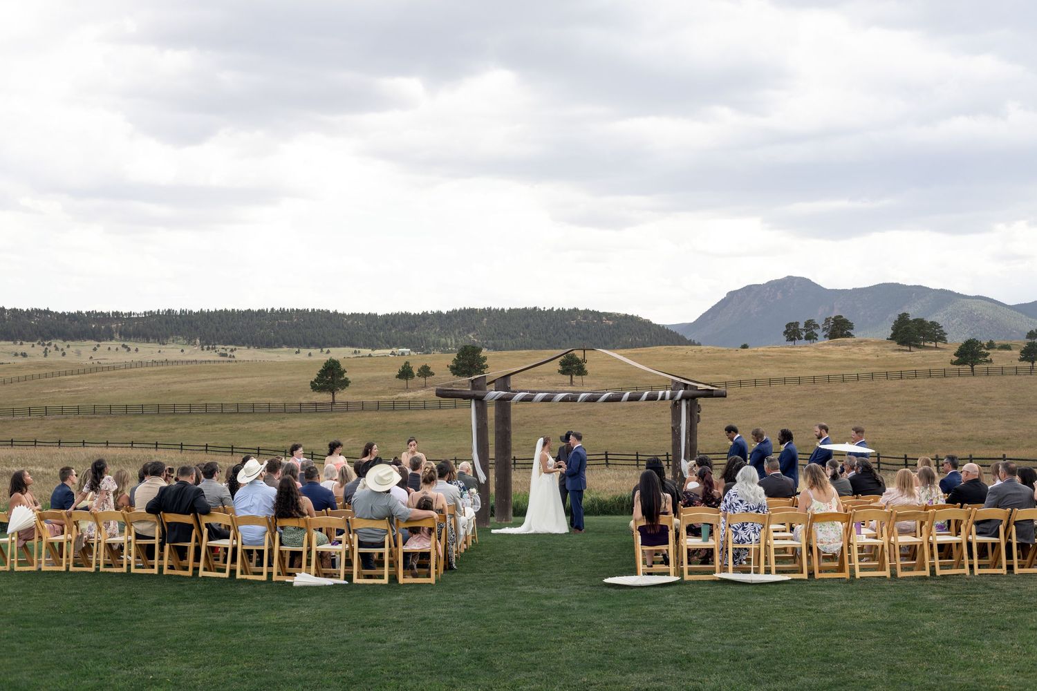 Outdoor wedding ceremony on lush green lawn with mountain views, guests seated in wooden chairs facing couple at rustic altar.
