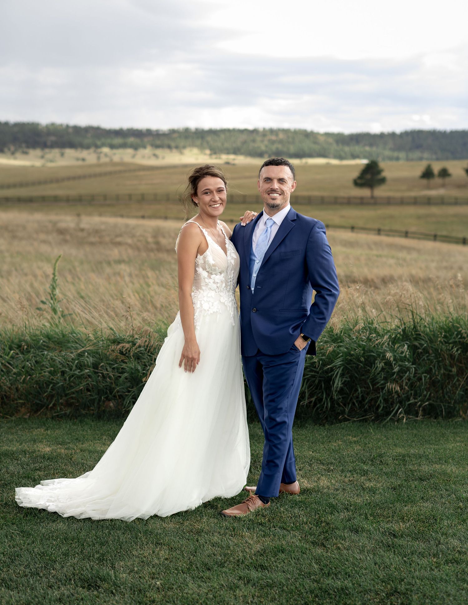 A smiling bride in a white lace gown and groom in a blue suit pose together in a scenic countryside setting.