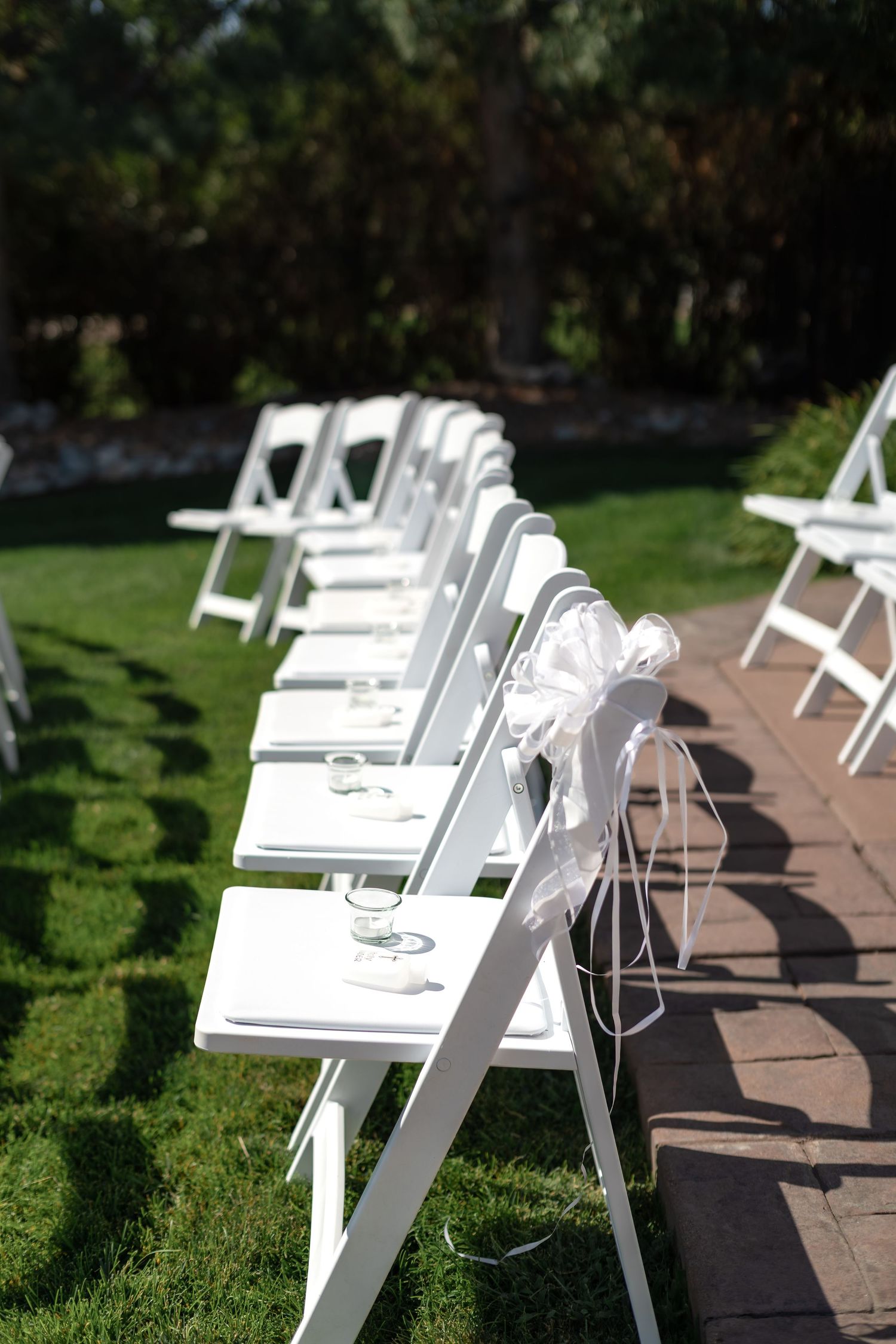 White folding chairs with ribbon bows line an outdoor wedding ceremony aisle on lush green grass.