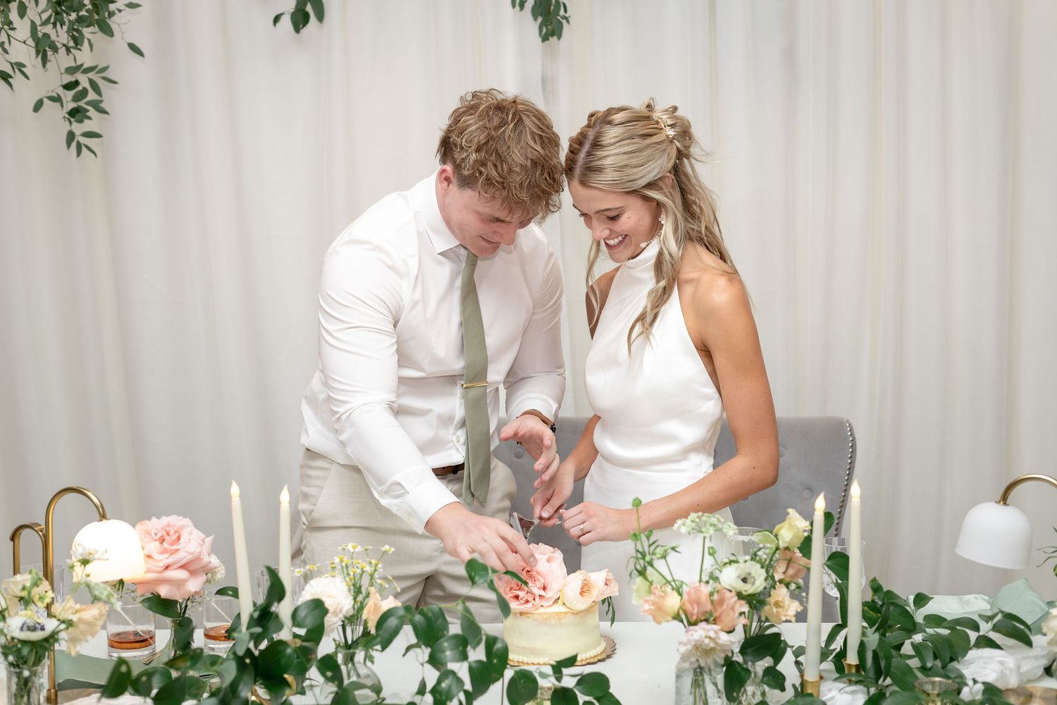 Bride and groom cutting wedding cake at elegant reception table with floral greenery and candles.