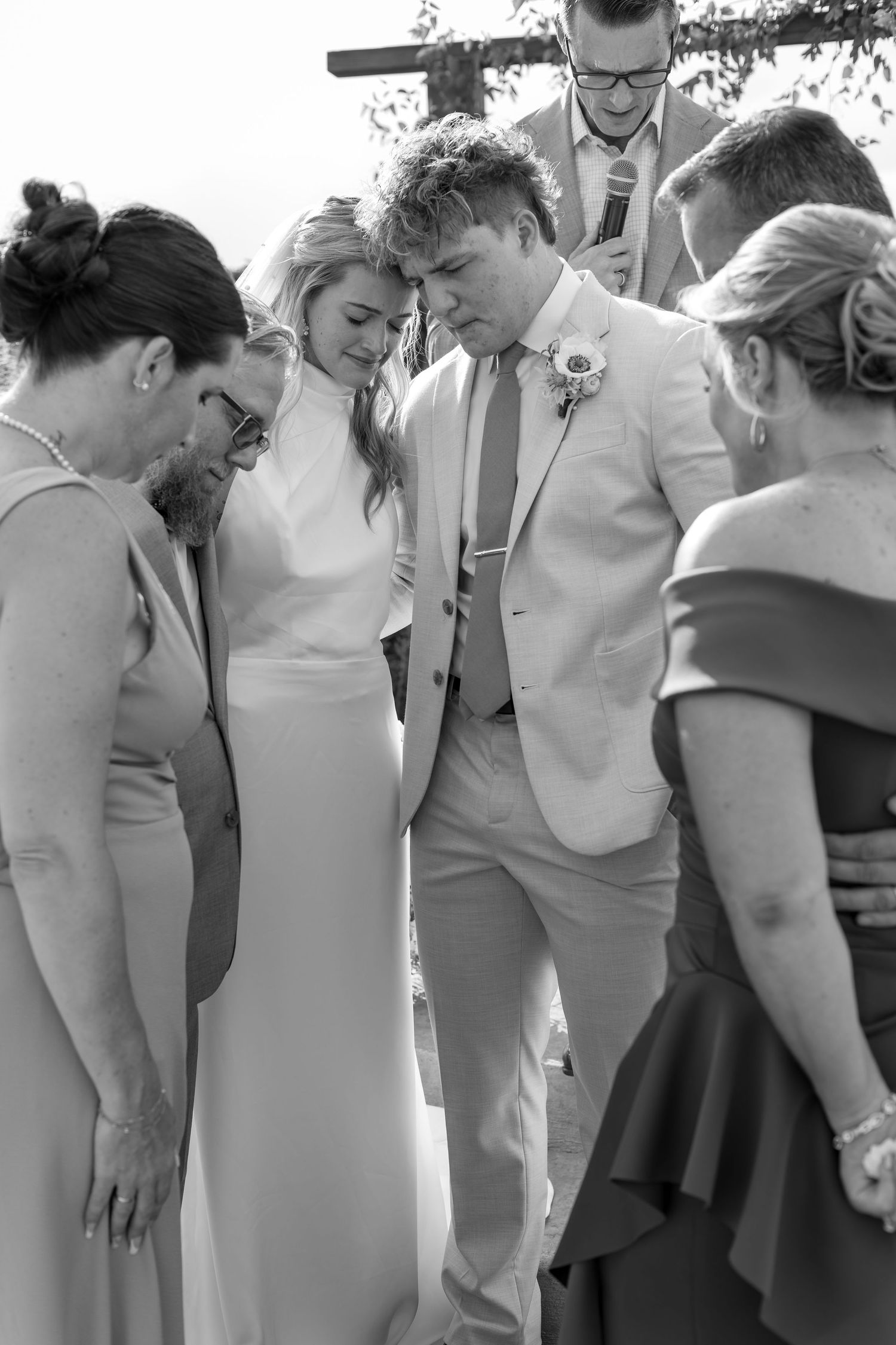 Black and white photo of wedding party gathered in prayer around bride and groom during ceremony.