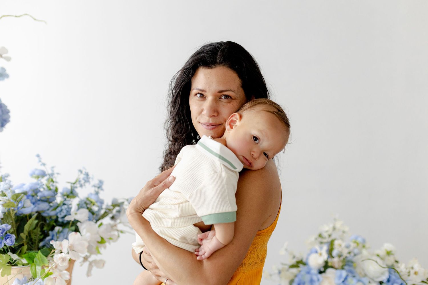 A mother in a yellow dress tenderly holds her smiling baby amid blue hydrangeas in a bright studio.