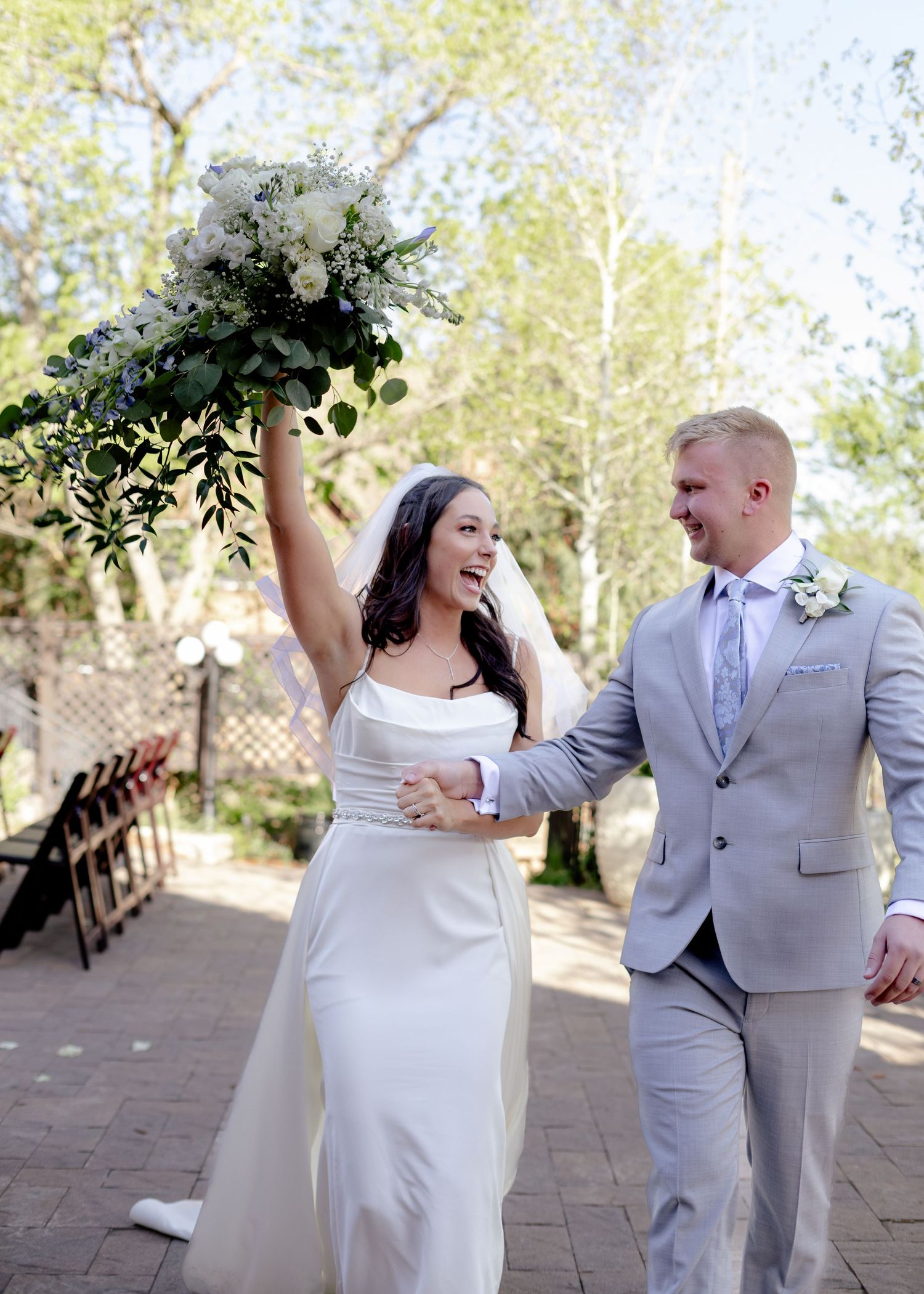 Joyful bride raises bouquet of white flowers while walking with groom after outdoor wedding ceremony.