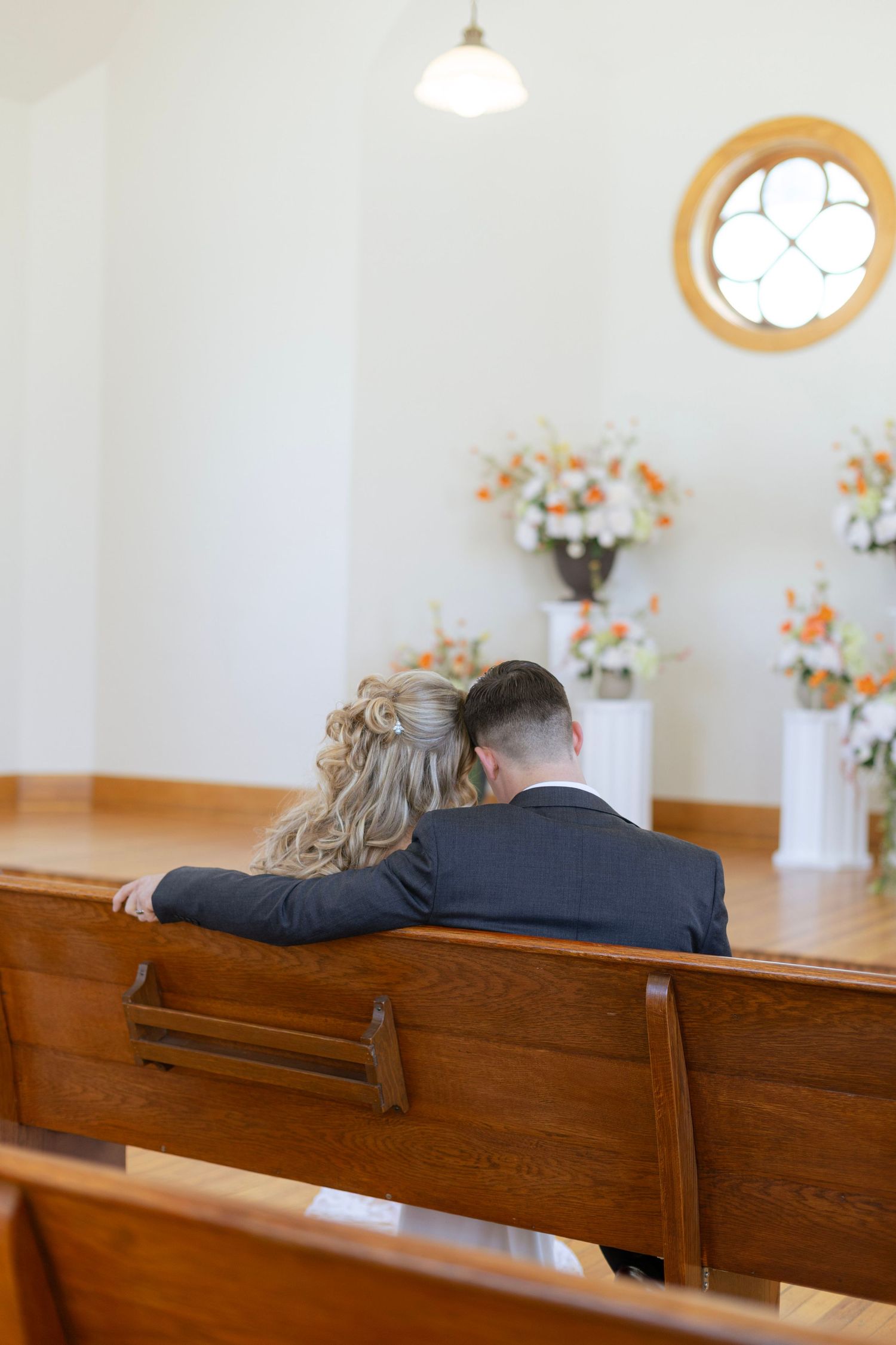 Bride and groom sit together on a wooden church pew, heads touching, facing floral altar arrangements.