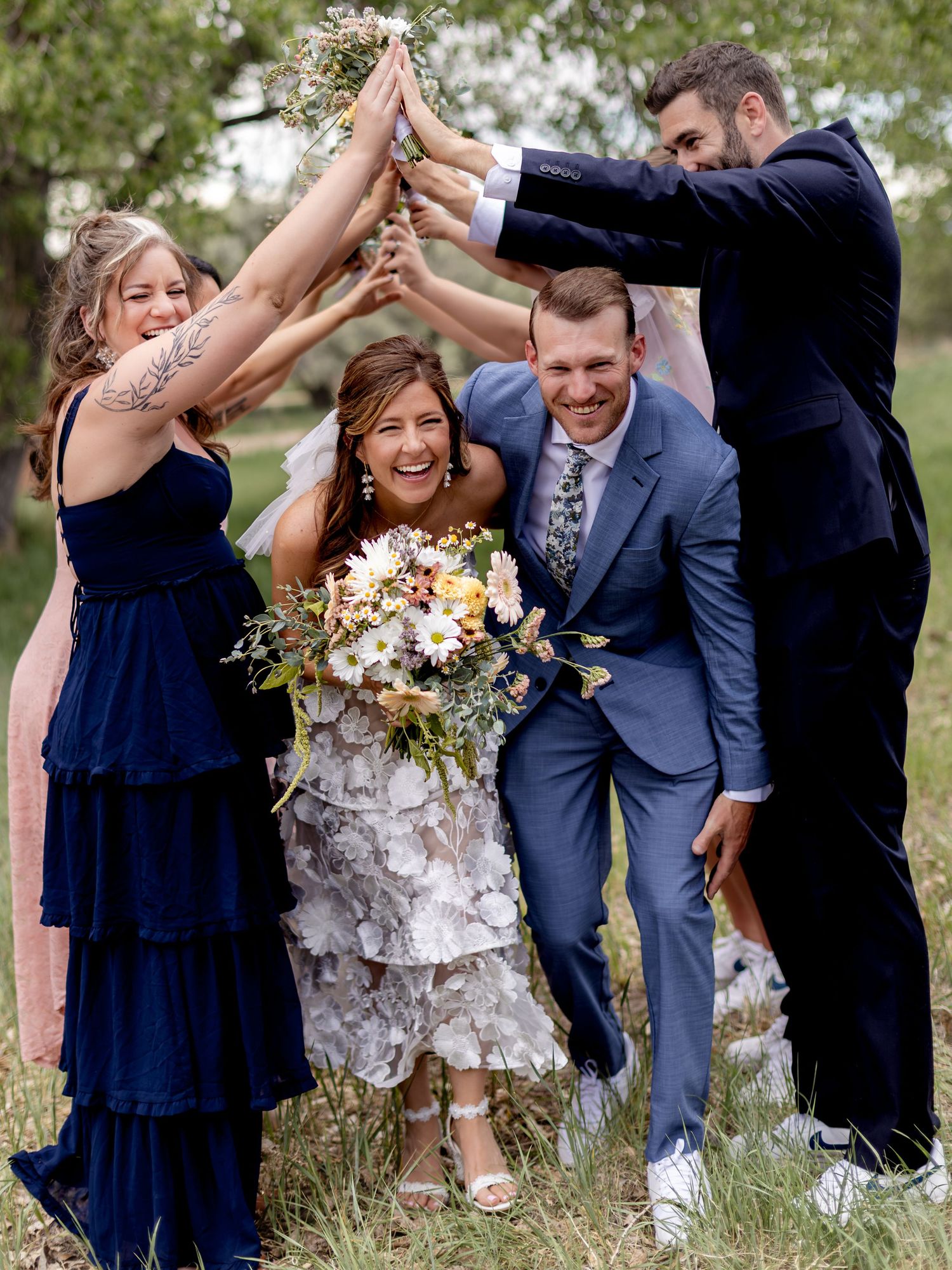 Bride and groom celebrate with wedding party raising hands in arch outdoors, bride holding wildflower bouquet.