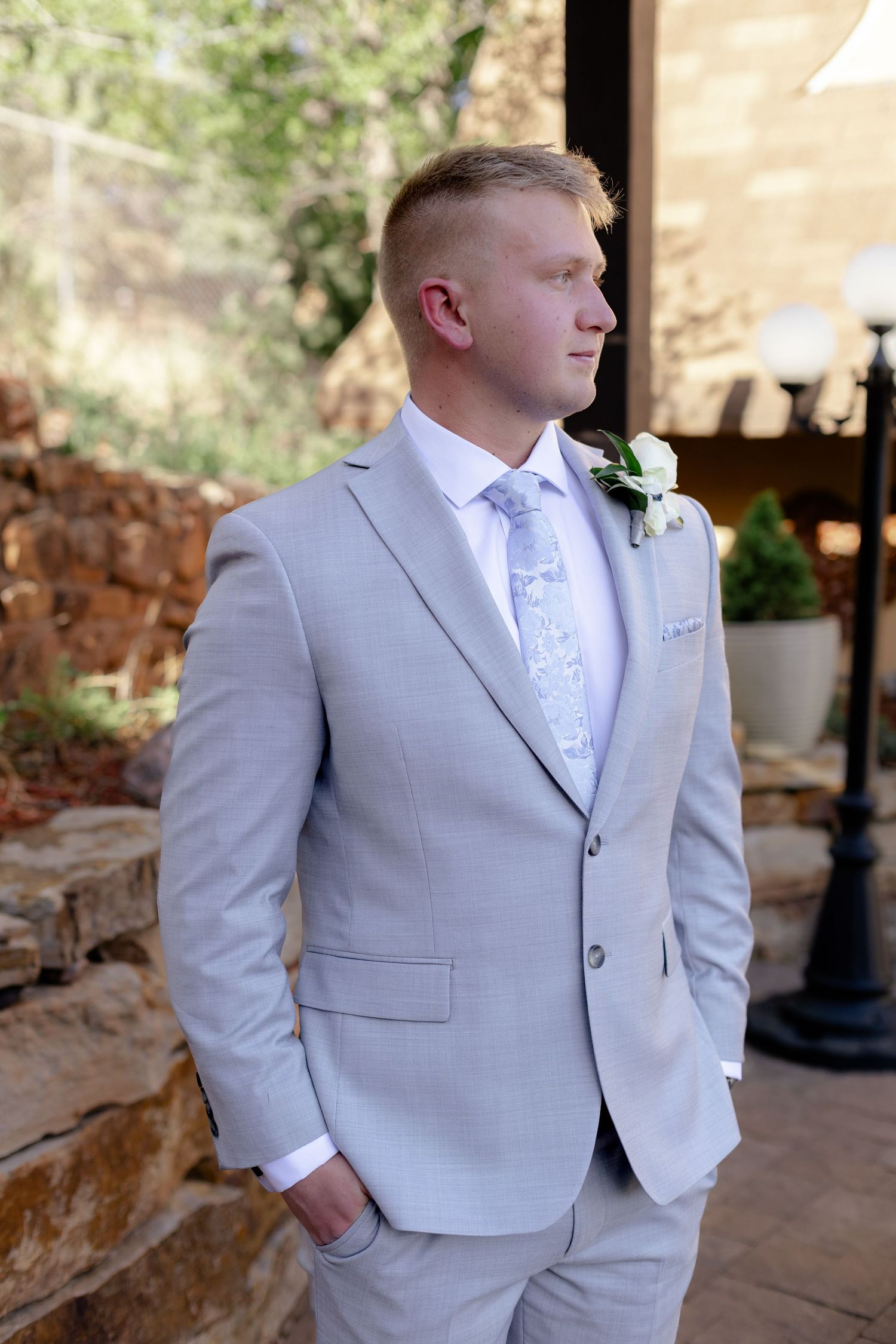 Groom in light gray suit with floral tie and white boutonniere poses outdoors at wedding venue.