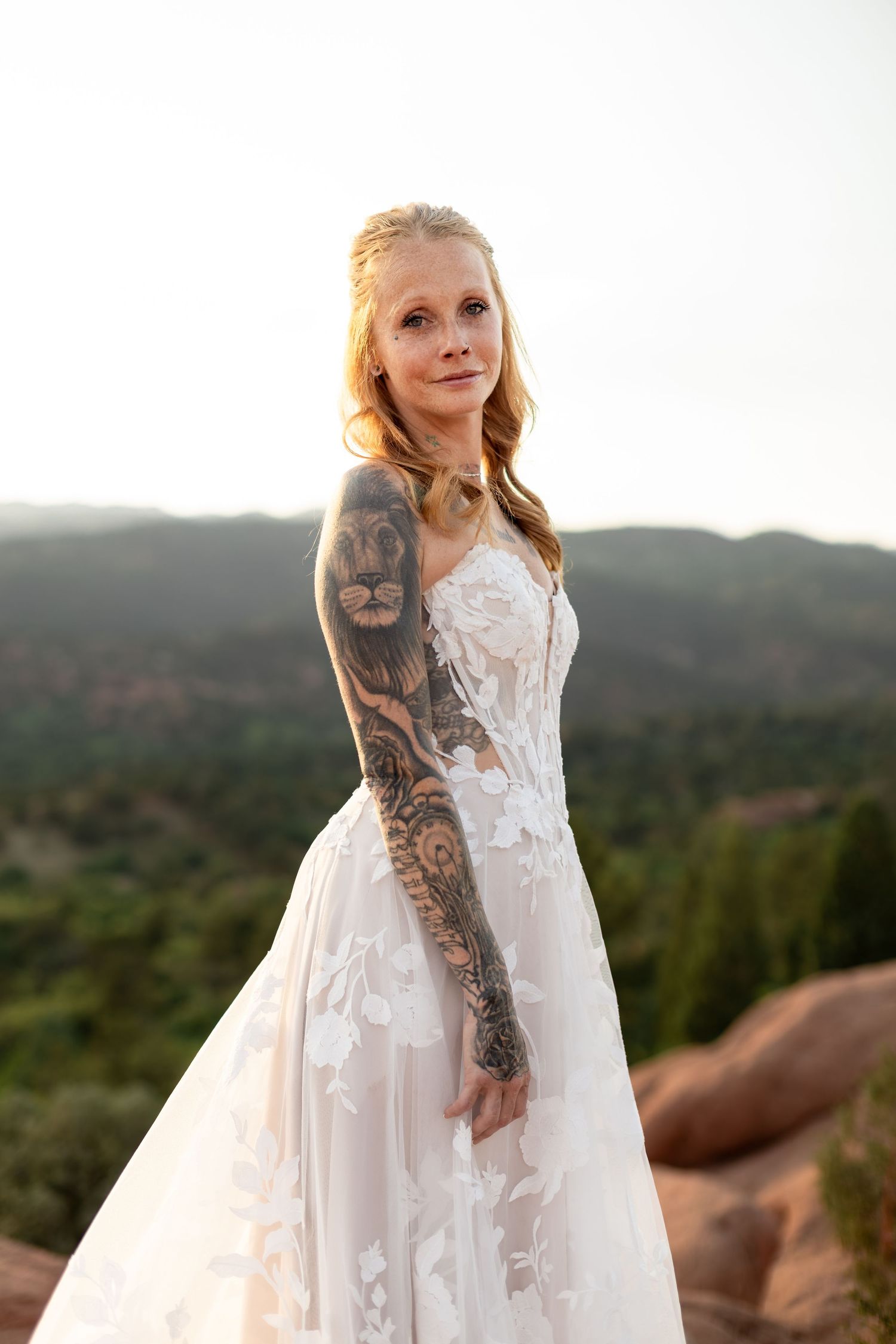 Tattooed bride in floral lace wedding gown posing at sunset with mountain landscape backdrop.