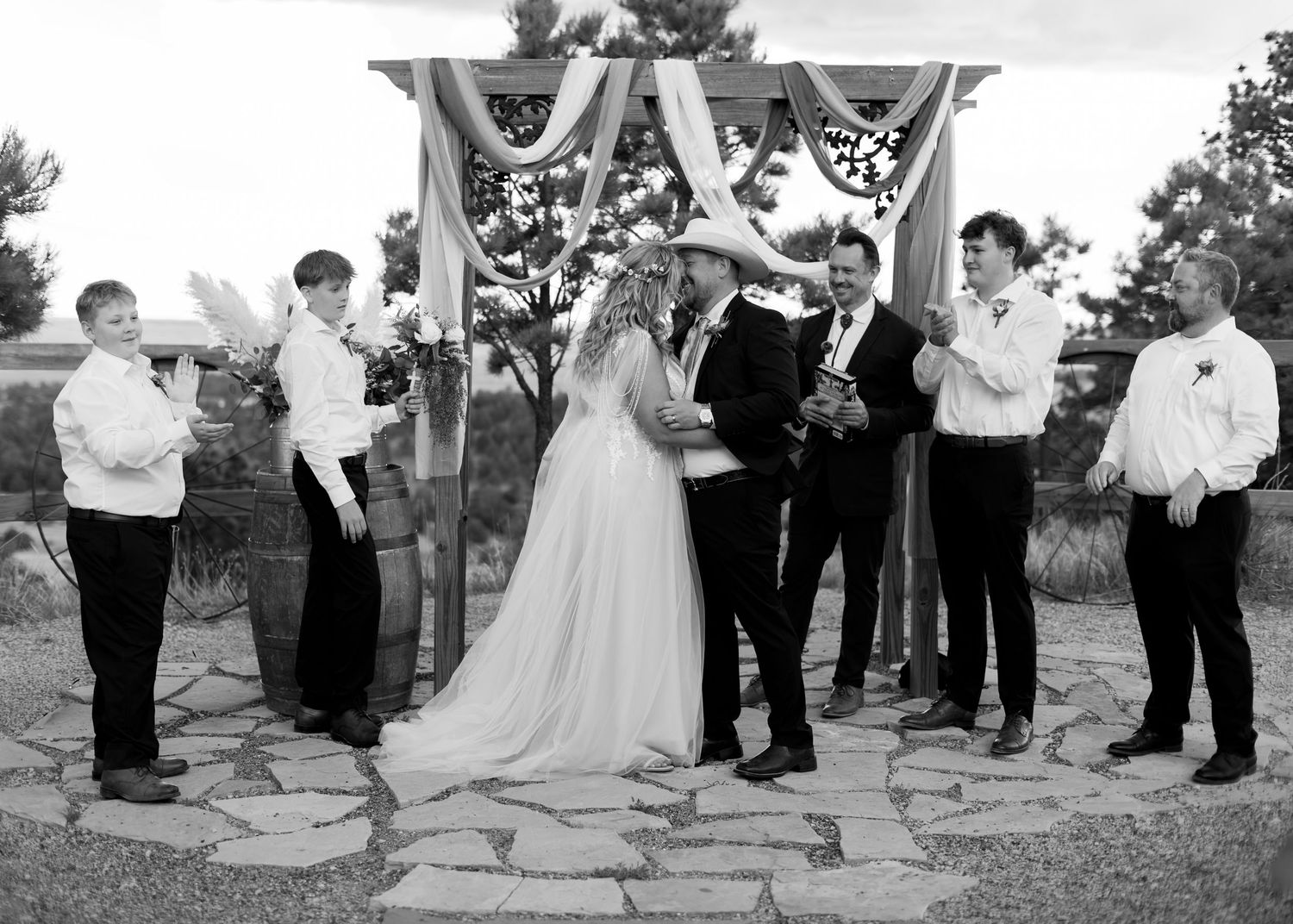 Wedding ceremony with bride and groom exchanging vows under a floral arch surrounded by groomsmen outdoors.