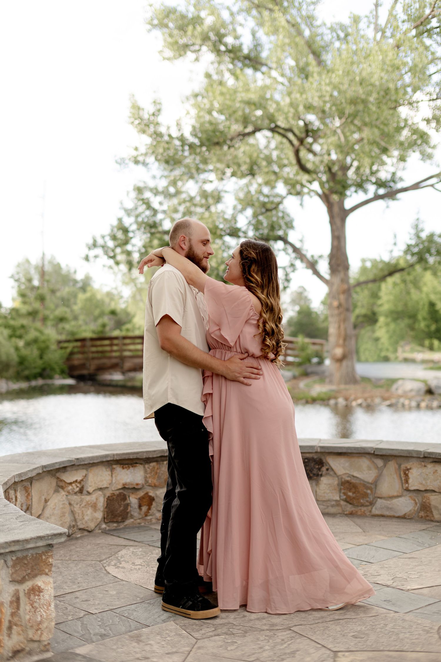 A couple embraces on a stone patio near a pond, woman in pink maxi dress, man in cream shirt, surrounded by lush trees.